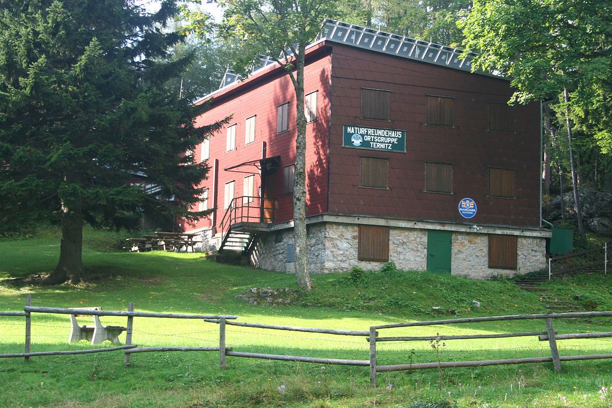 Červená budova s dreveným obložením, obklopená stromami a lúkou, s nápisom "Naturfreundehaus Ortsgruppe Ternitz".