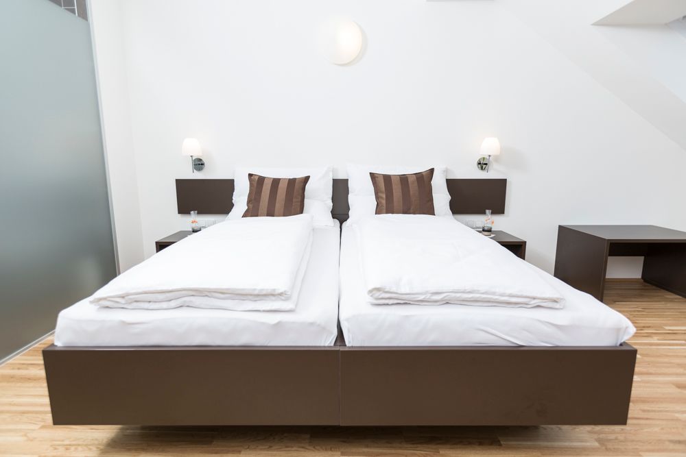 Moderná izba pre hostí s dvoma samostatnými posteľami, bielymi perinami a hnedými vankúšmi, drevenou podlahou a nočnými stolíkmi s lampami.