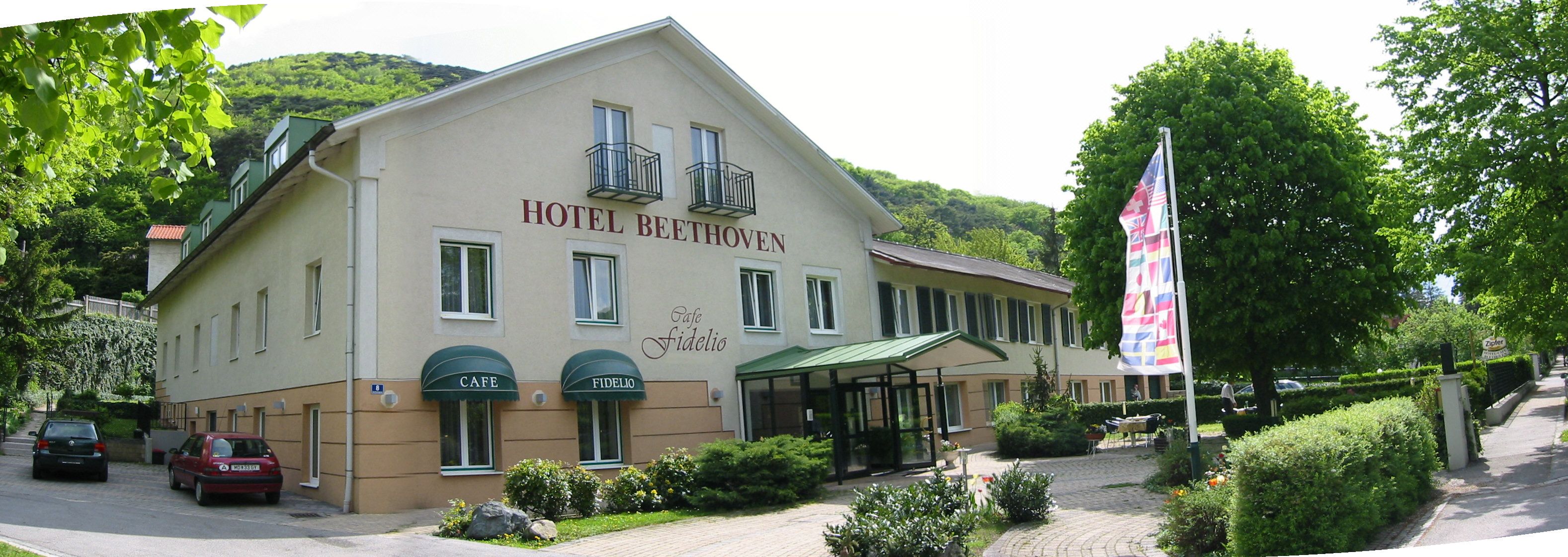 Exteriérový pohľad na hotel Beethoven s kaviarňou Fidelio, obklopený stromami a vlajkami.