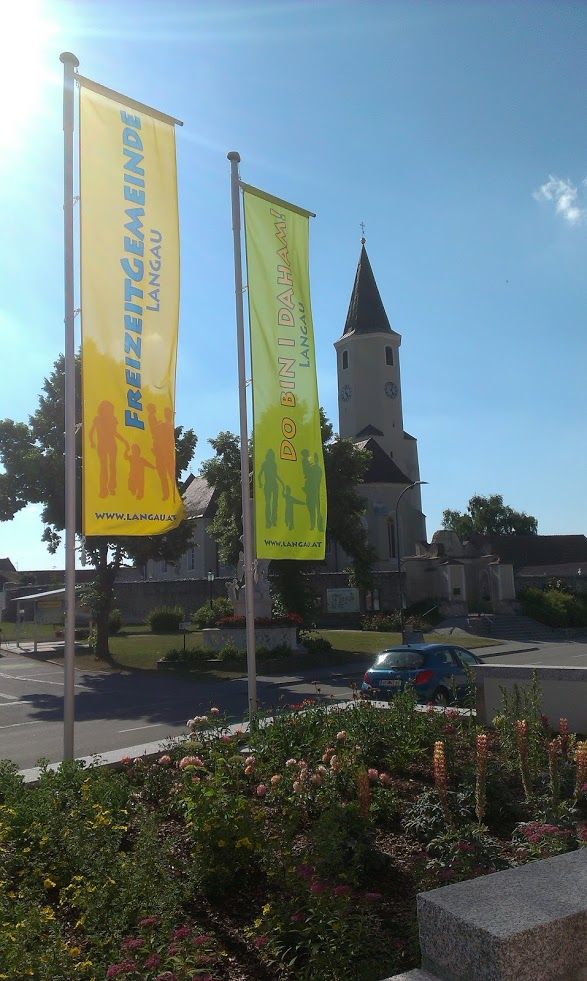 Dve farebné vlajky s nápismi "Freizeitgemeinde Langau" a "Do bin i daham" pred kostolom.