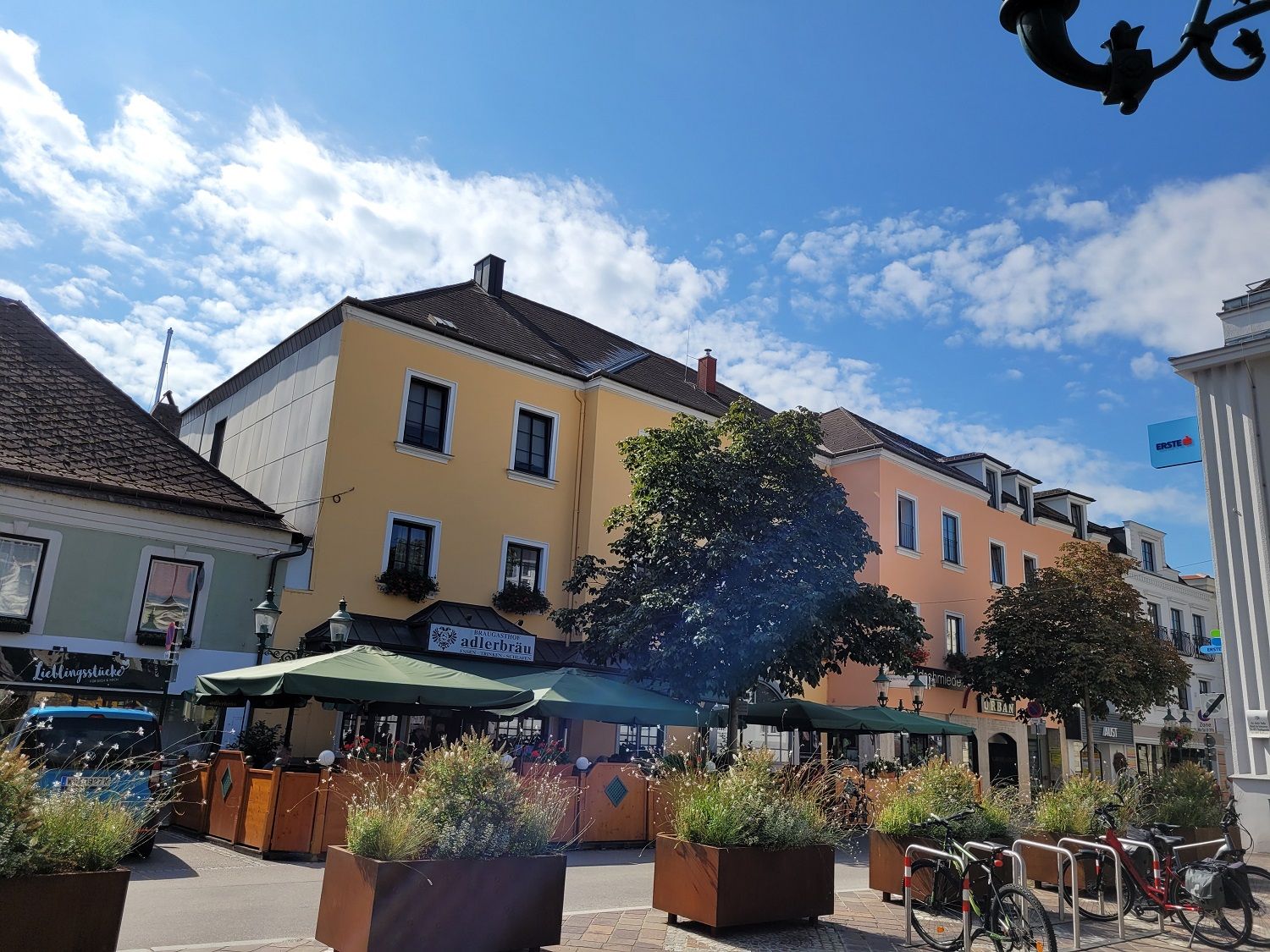 Scéna ulice so žltou budovou, kaviarňou so slnečníkmi a bicyklami v popredí.