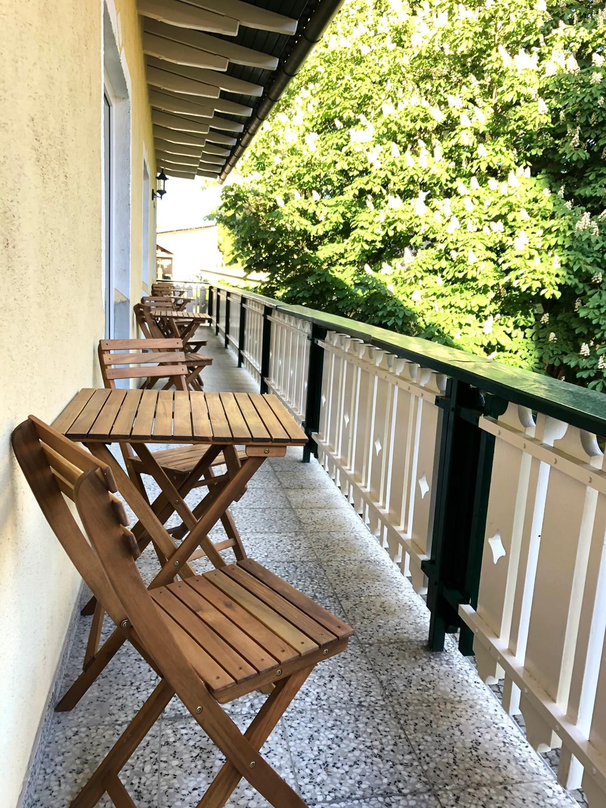 Drevený balkón so stolom a stoličkami, vedľa neho zelené stromy.