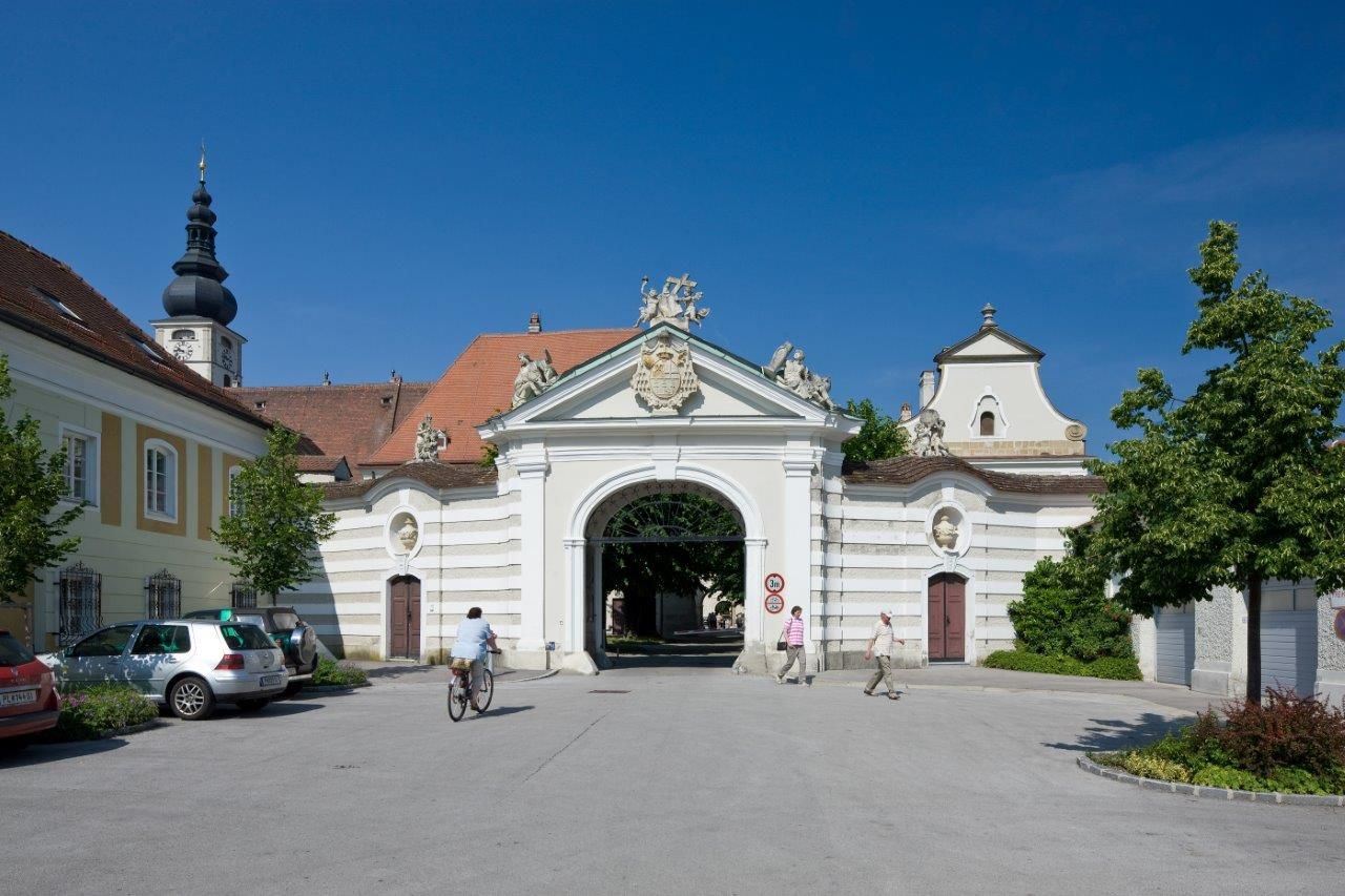 Historická budova diecézy s oblúkom a kostolnou vežou v pozadí.