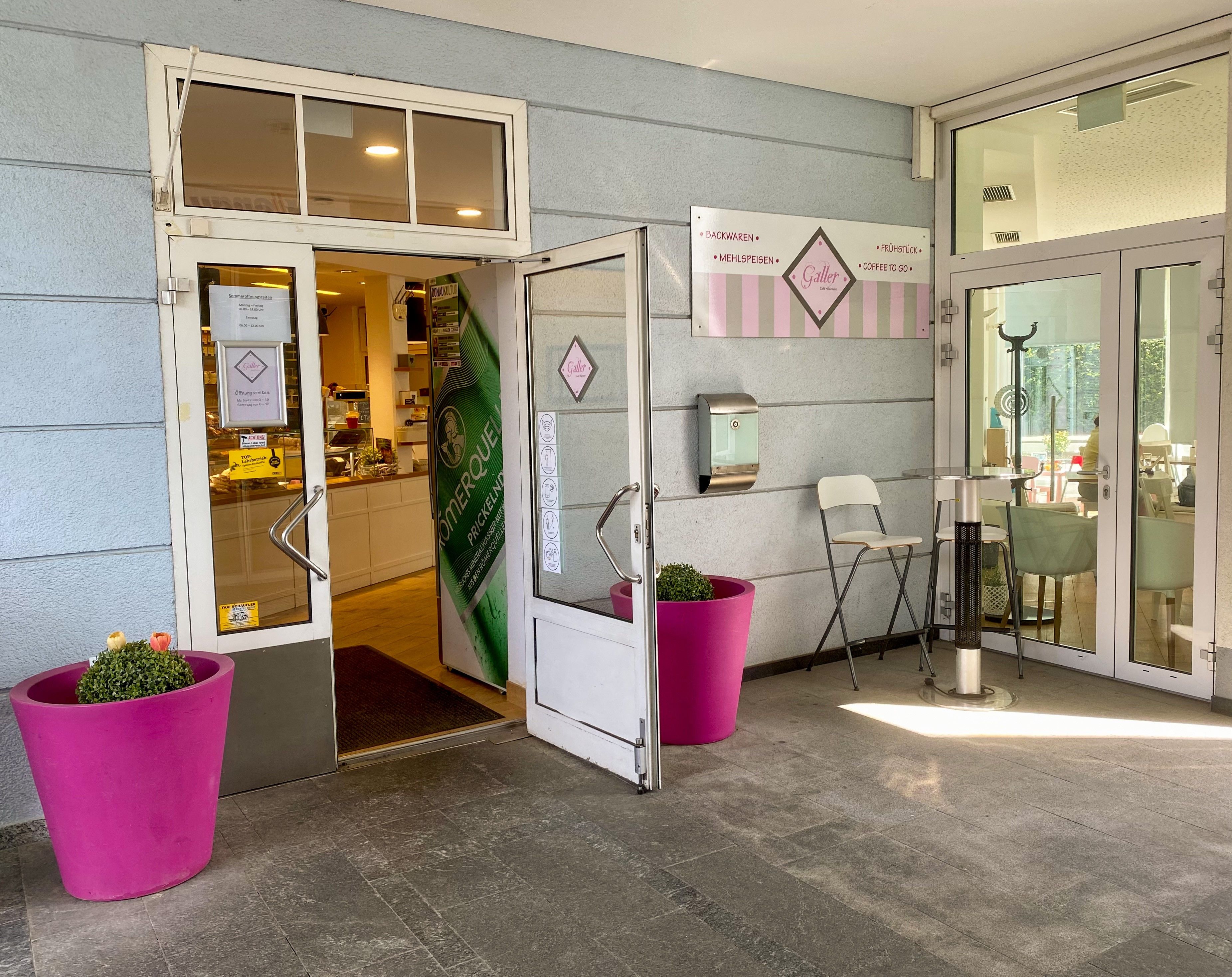 Vchod do kaviarne a vpravo ružové kvetináče pred svetlomodrým domom 