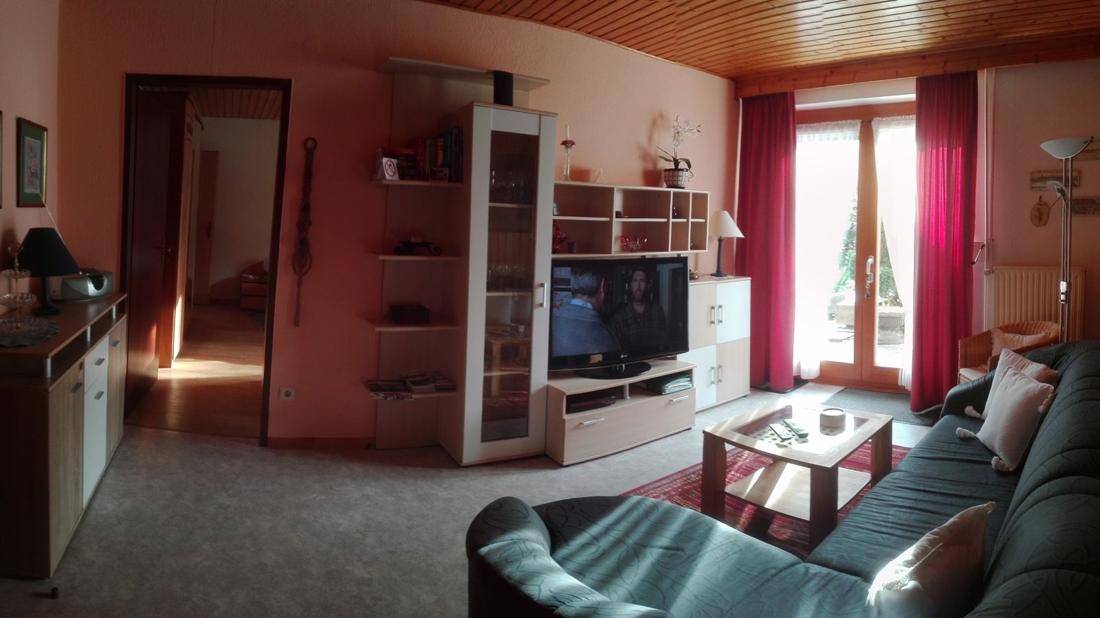Útulná obývacia izba s pohovkou, TV, policou a sklenenými dverami s červenými závesmi.