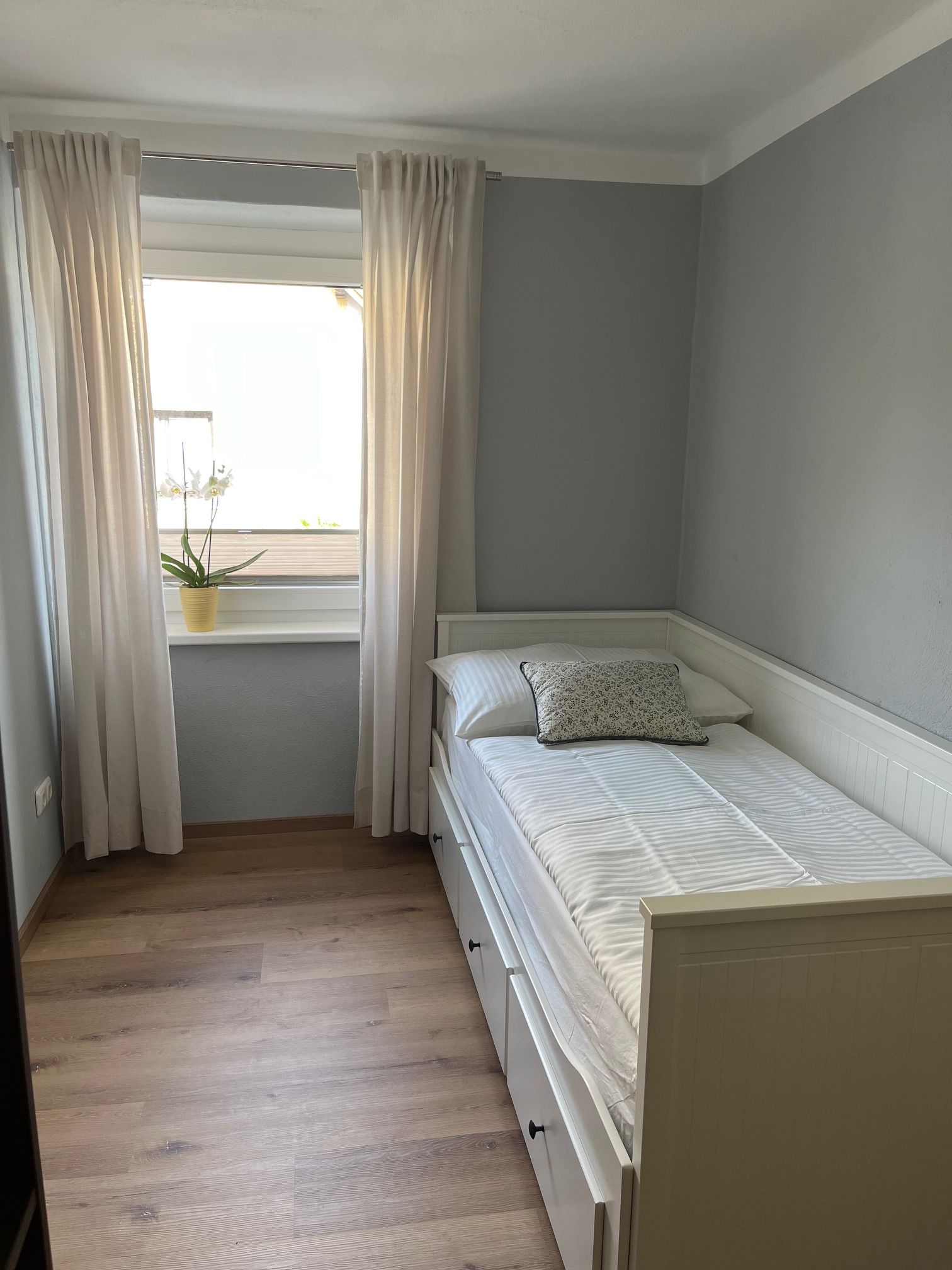 Malá spálňa s jednolôžkom, bielymi závesmi a rastlinou na parapete.