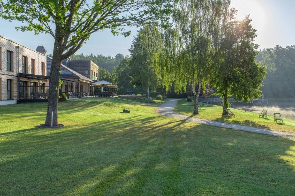 Zelená lúka so stromami, budovami a ležadlami na brehu jazera v slnečnom svetle.