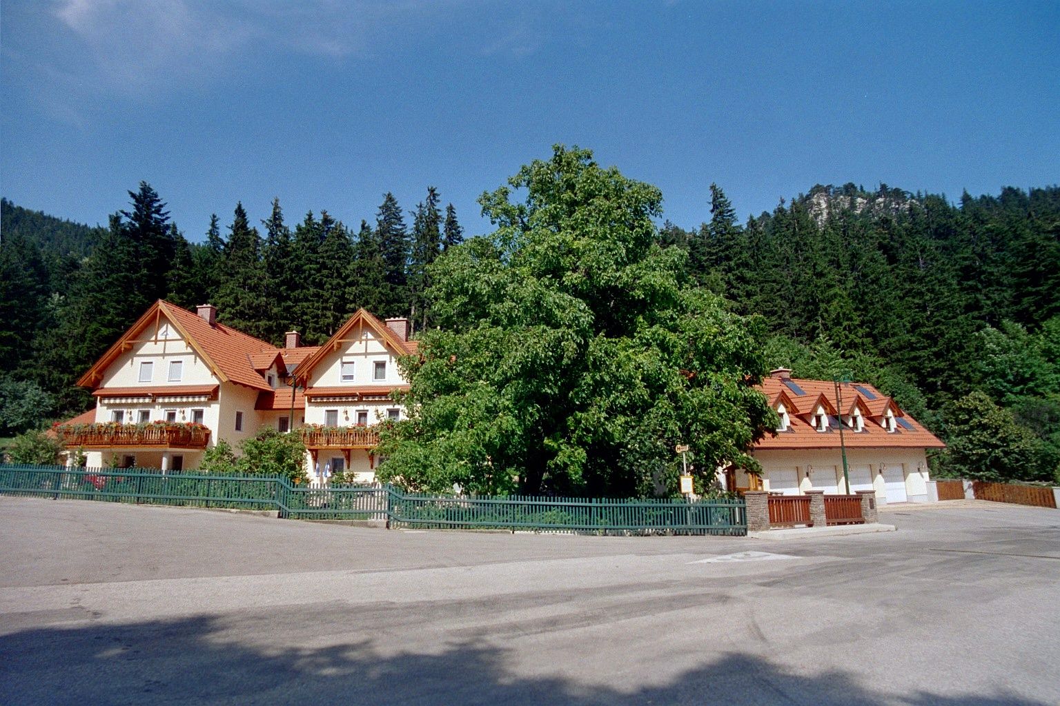 Veľký dom s červenými strechami a balkónmi, obklopený stromami a lesom v pozadí.