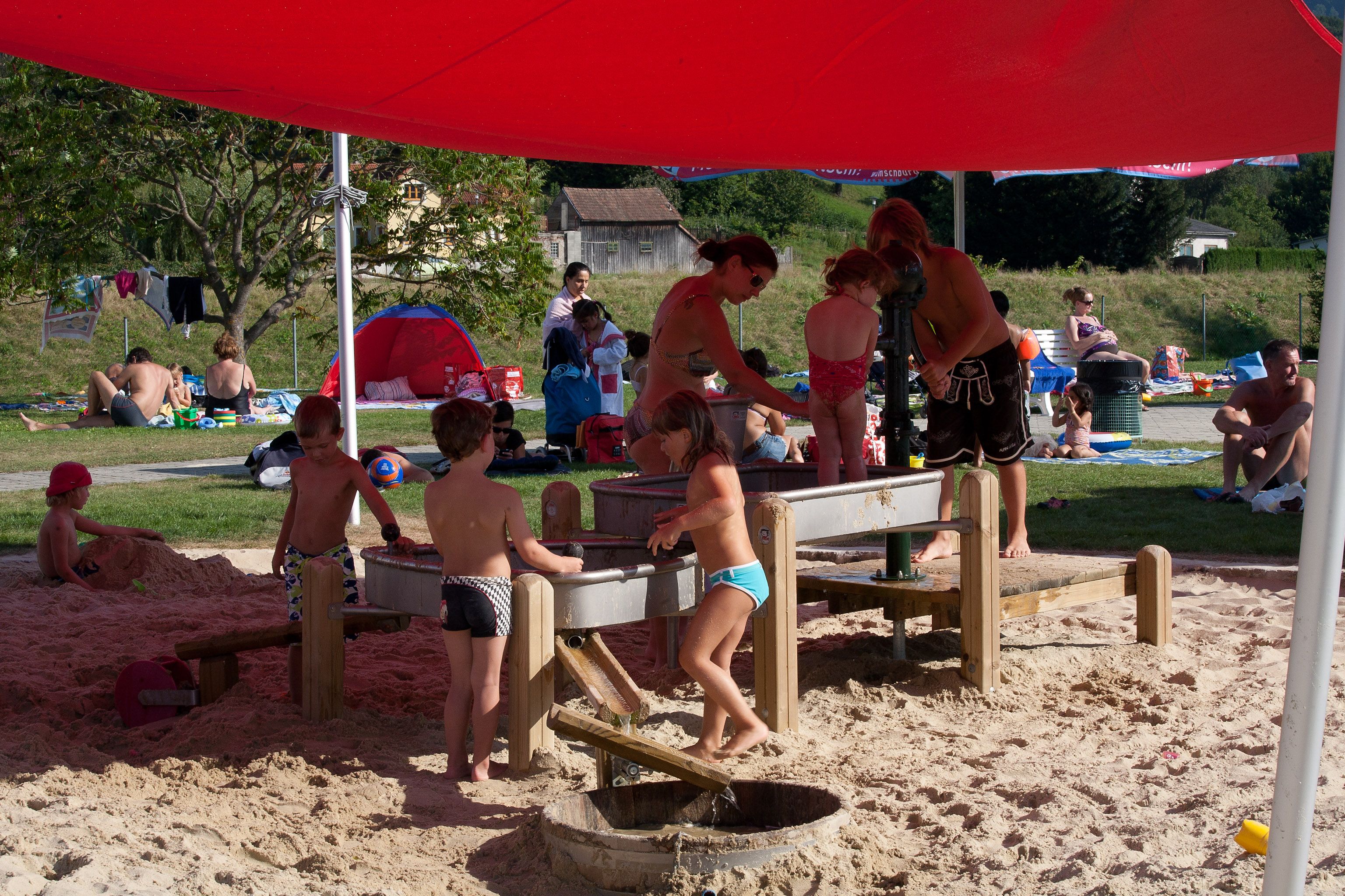 Deti sa hrajú v piesku pod červenou markízou vo Wilhelmsburg Parkbad.