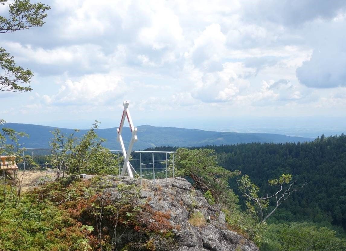 Pohľad na horu s bielym rámom v tvare kríža a výhľadom na zalesnené kopce pod zamračenou oblohou.