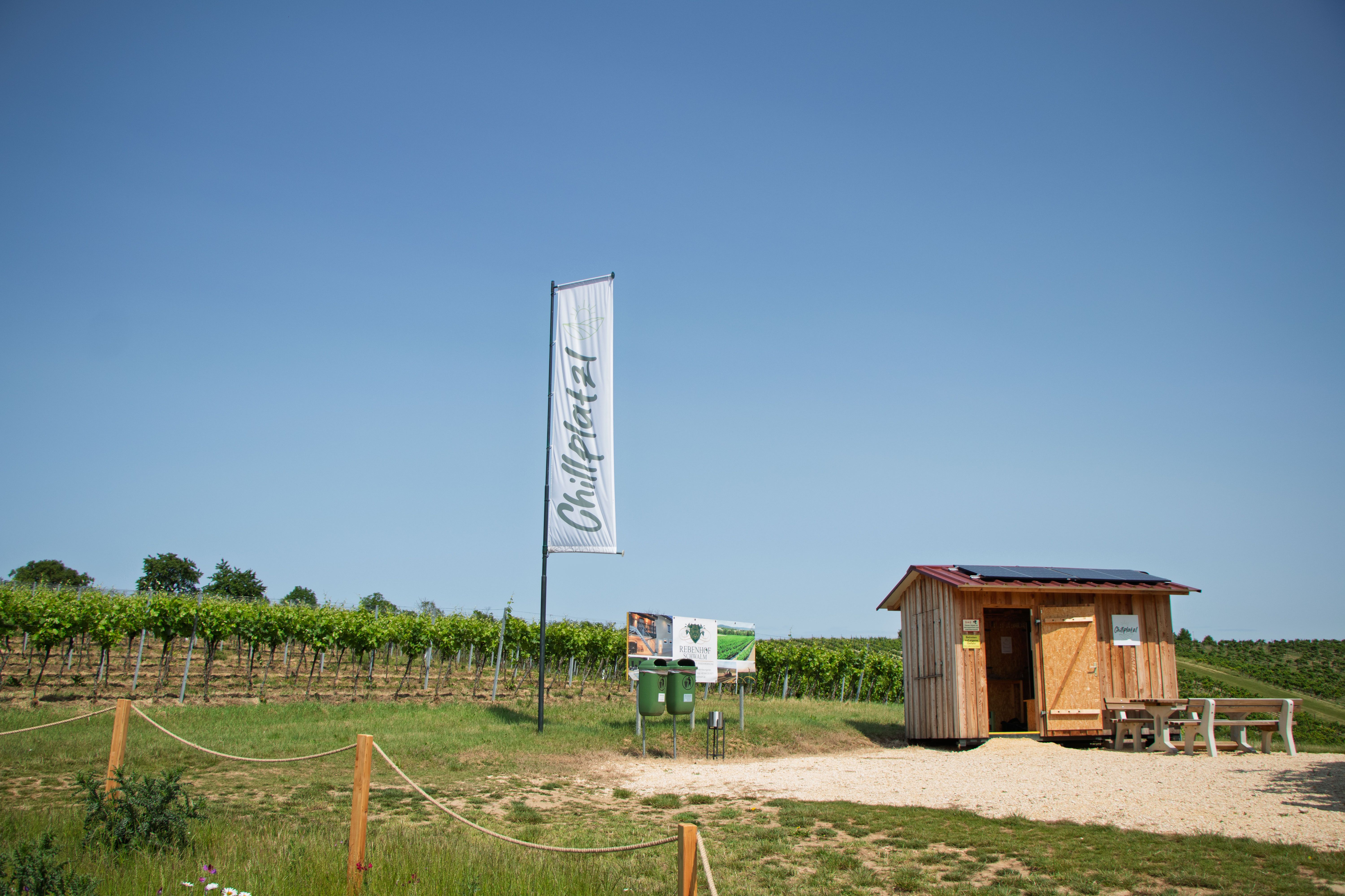 Malá drevená búdka vedľa vinice s tabuľkou a vlajkou s nápisom „Chillplatz“.