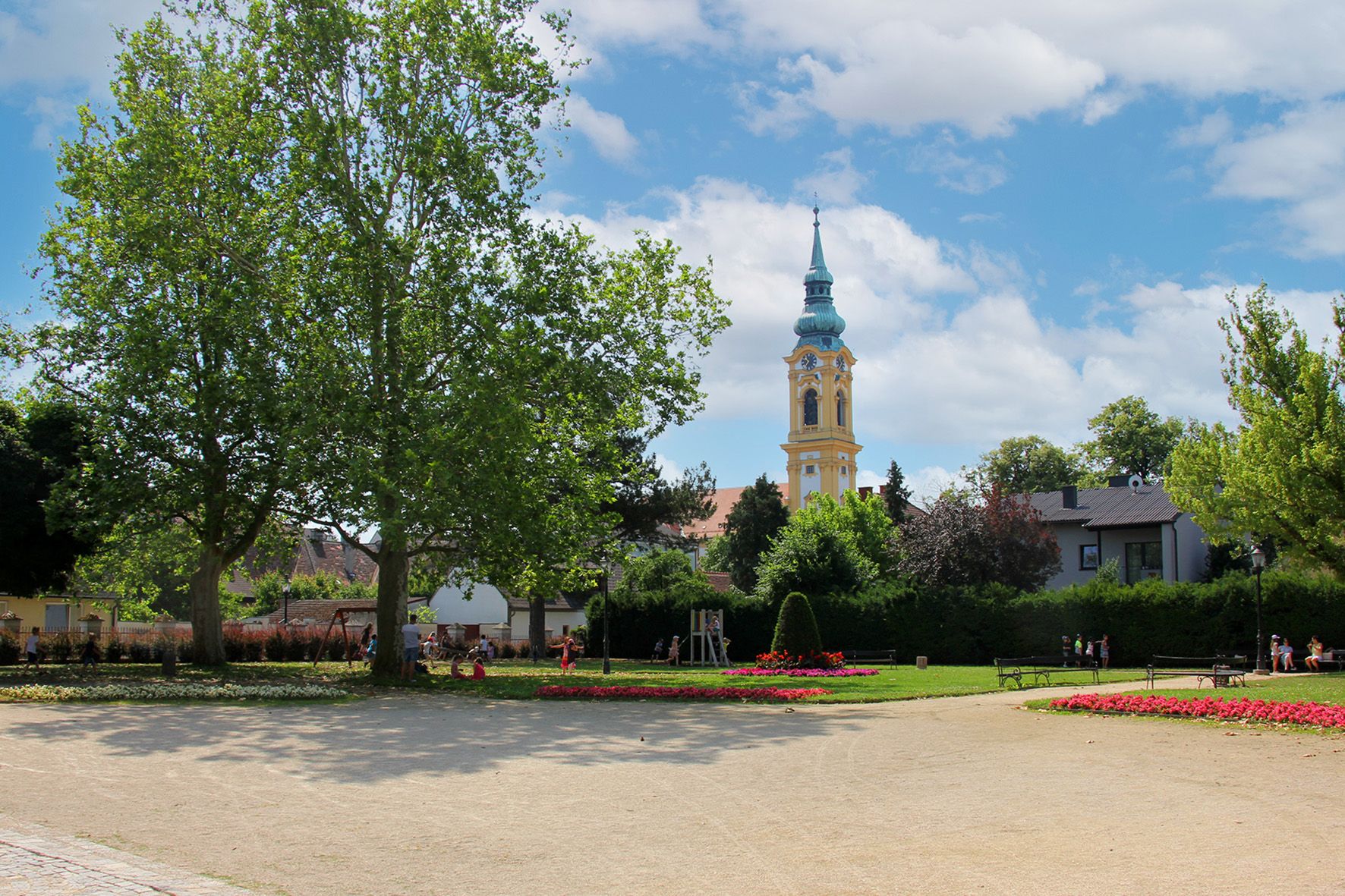 Park so stromami, kvetinovými záhonmi a kostolnou vežou v pozadí.