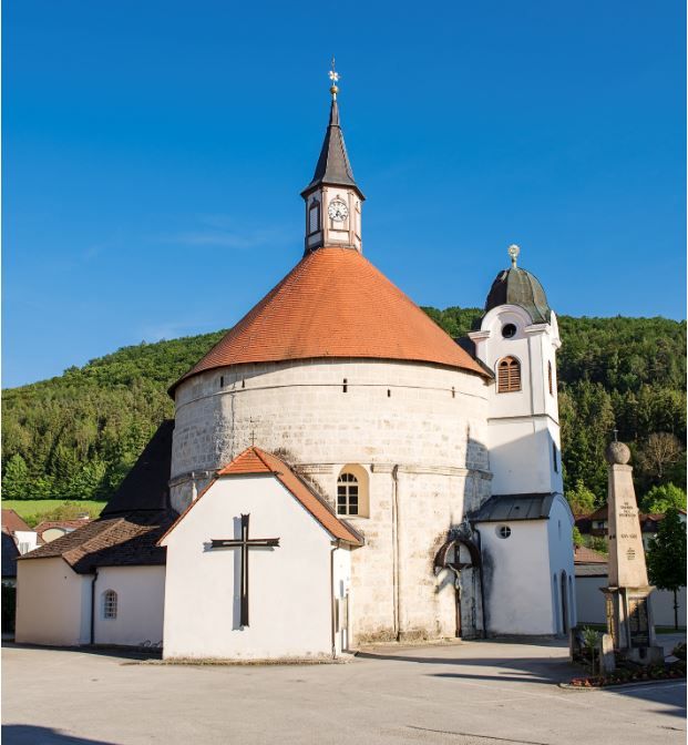 Farský kostol v Scheiblingkirchene s okrúhlou vežou a červenou strechou na pozadí modrej oblohy.
