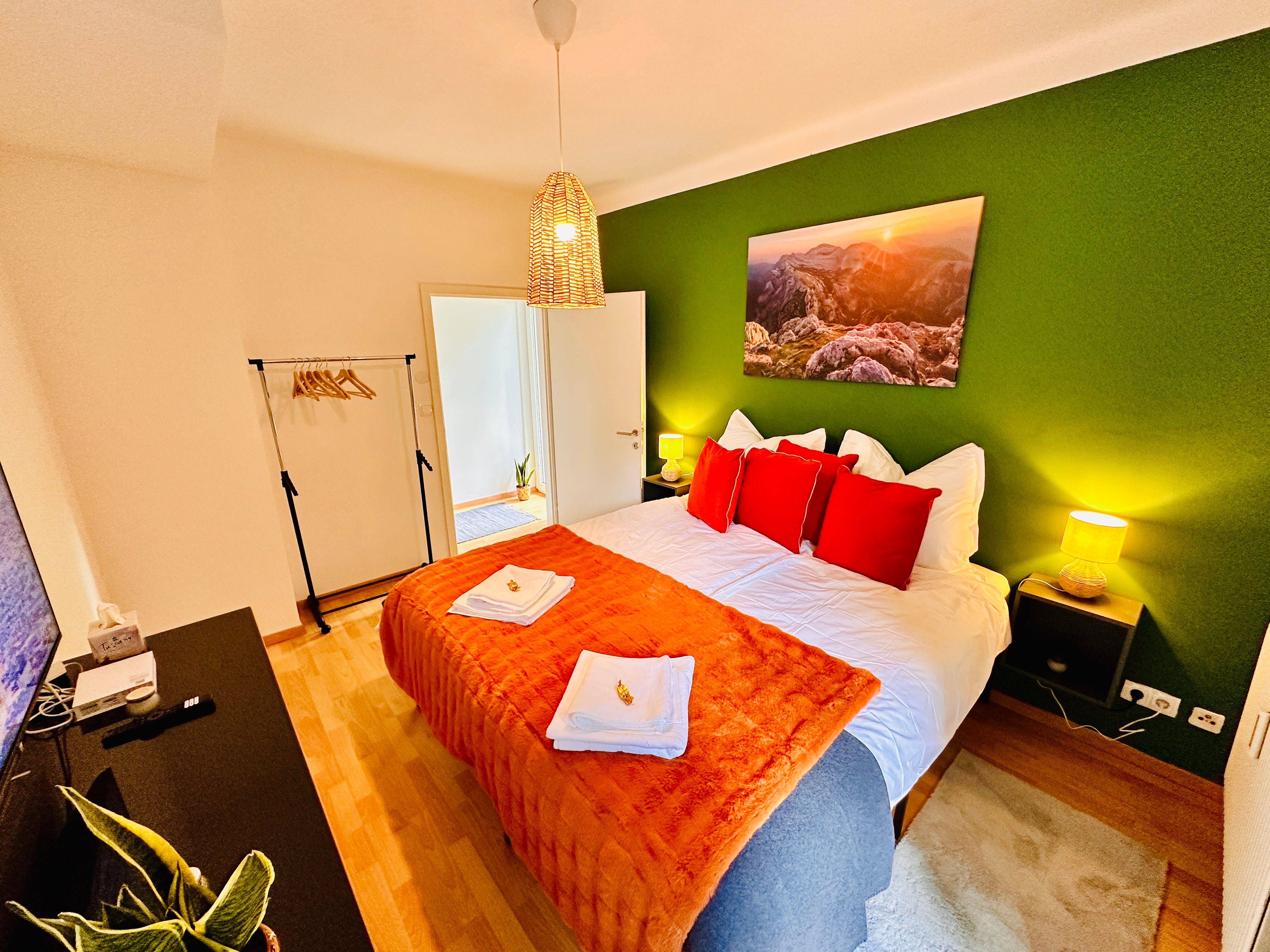 Útulná spálňa so zelenou stenou, manželskou posteľou s červenými vankúšmi, nočnými stolíkmi a lampou.