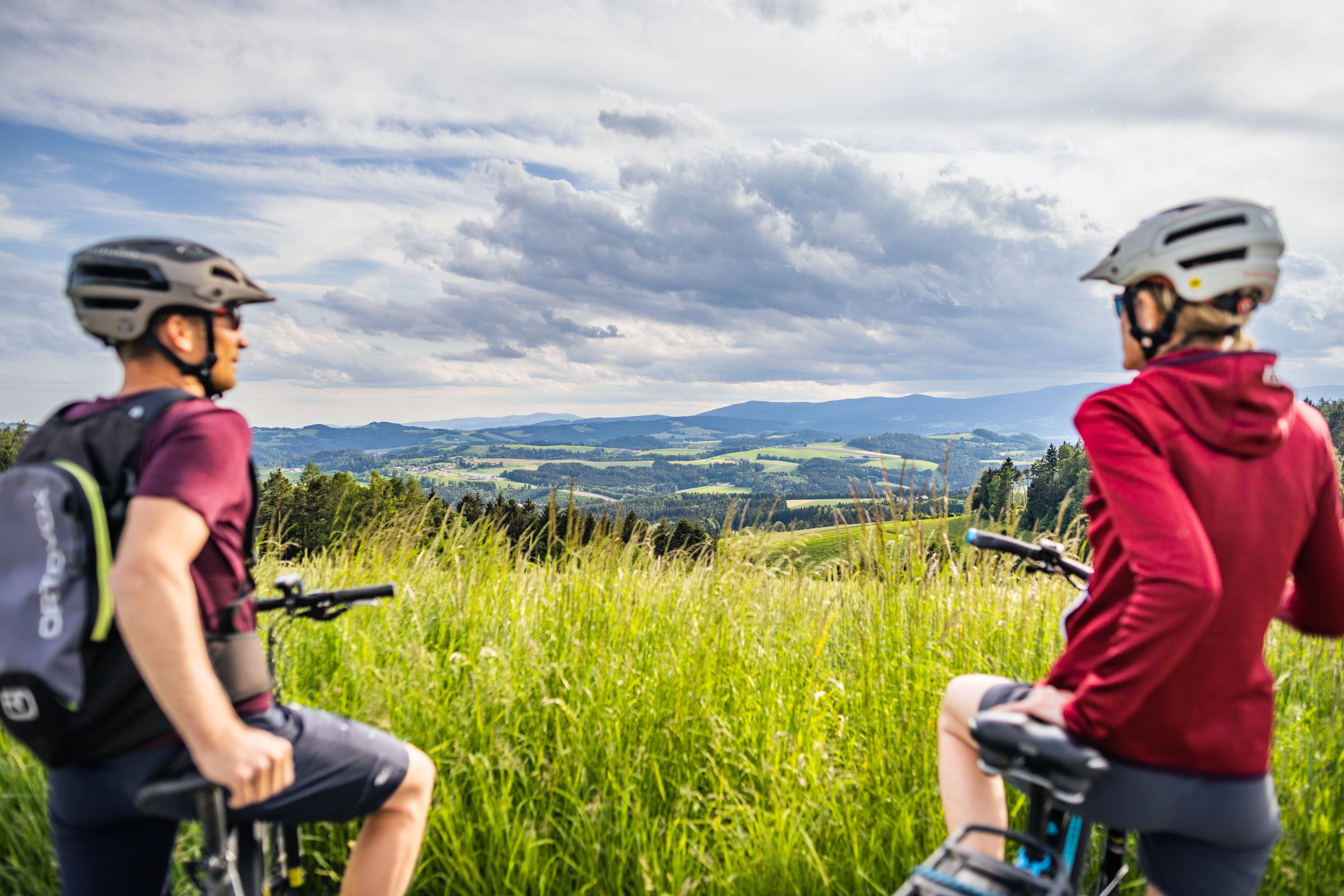 Dvaja cyklisti s prilbami sa pozerajú na kopcovitú krajinu lúk a lesov pod zamračenou oblohou.
