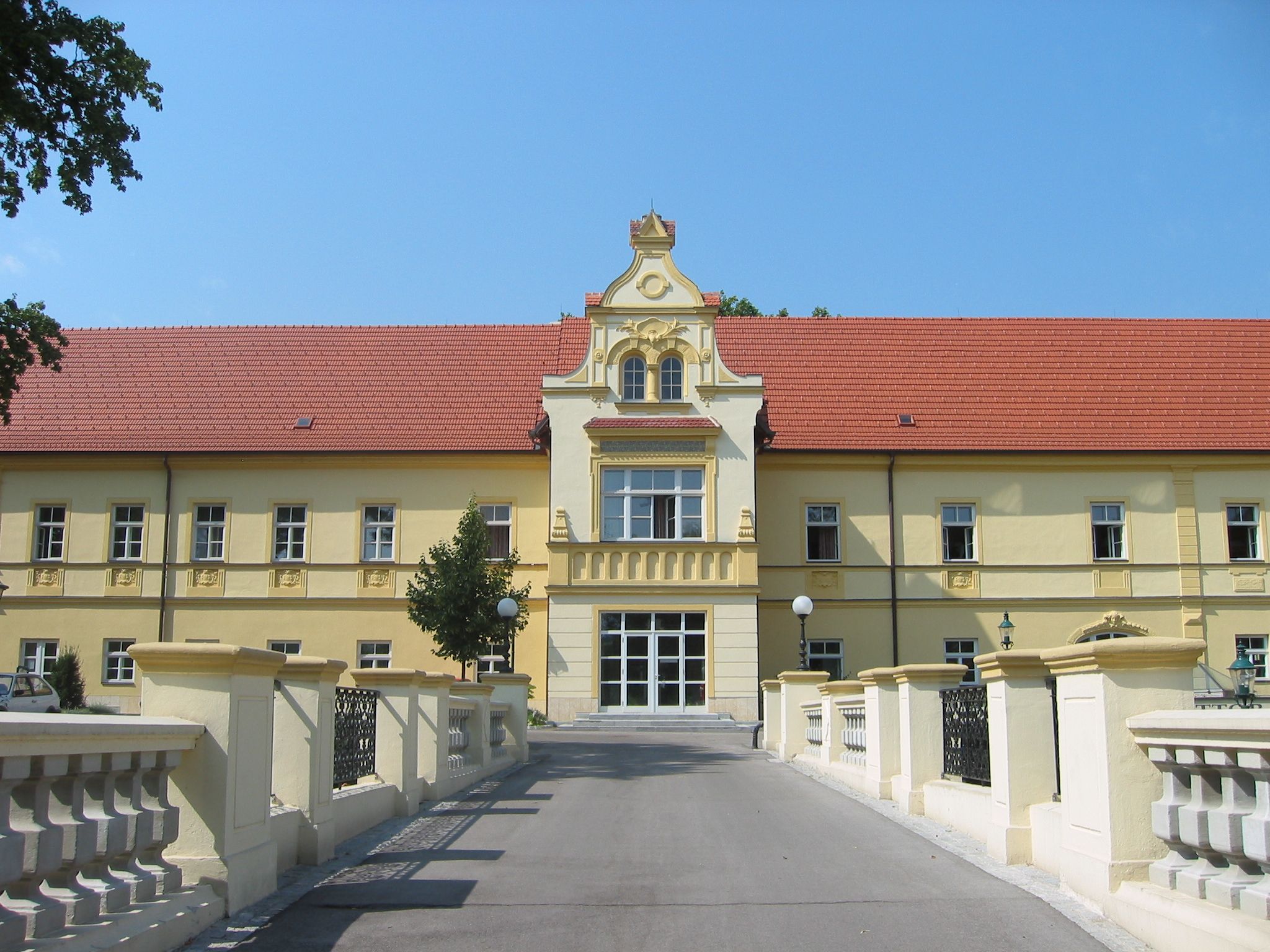 Žltá budova s červenou strechou a ozdobnou fasádou, obklopená mostom a stromami.