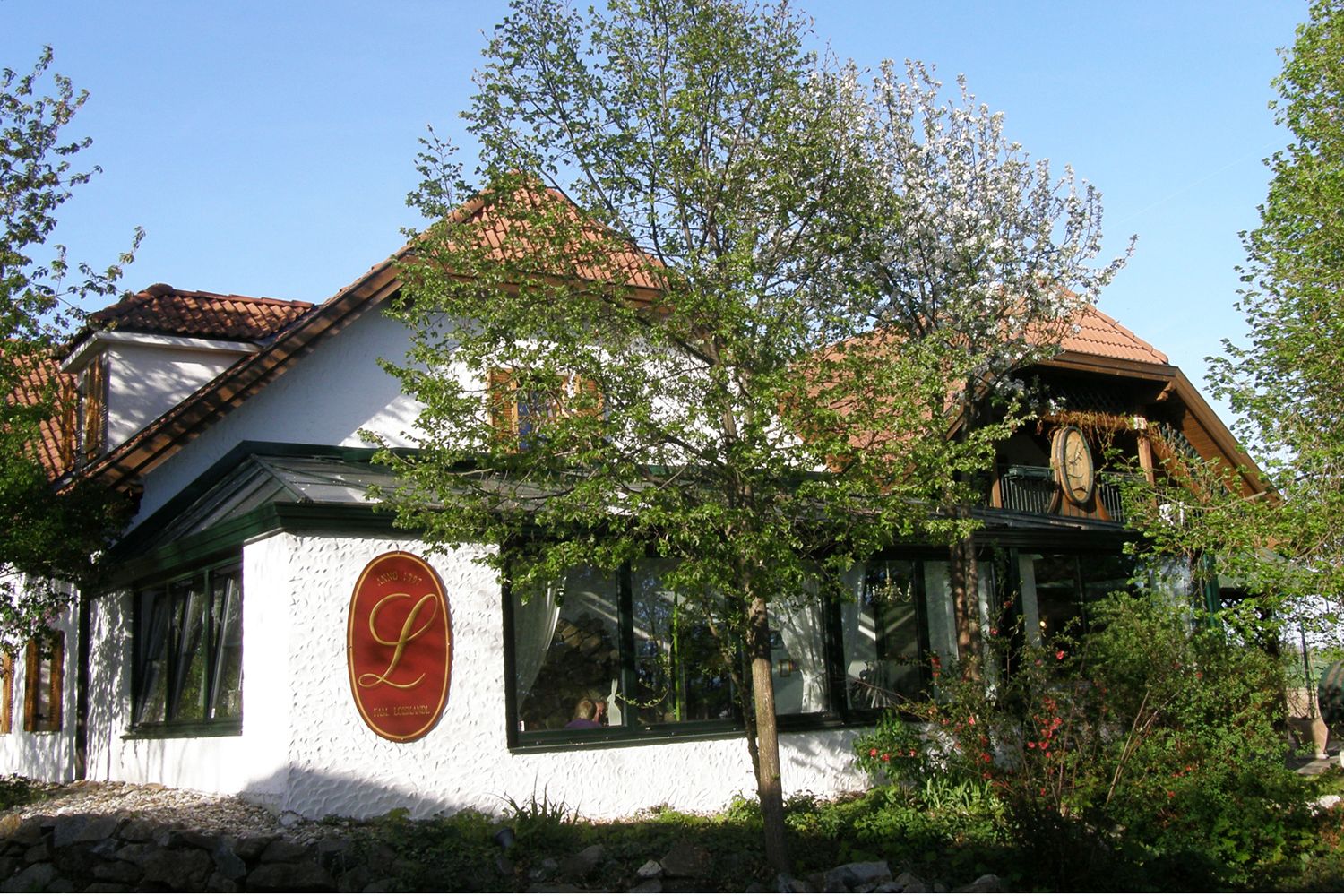 Pohľad zvonka na tradičnú budovu s červenými škridlami a veľkým logom L na stene, obklopenú stromami a rastlinami.