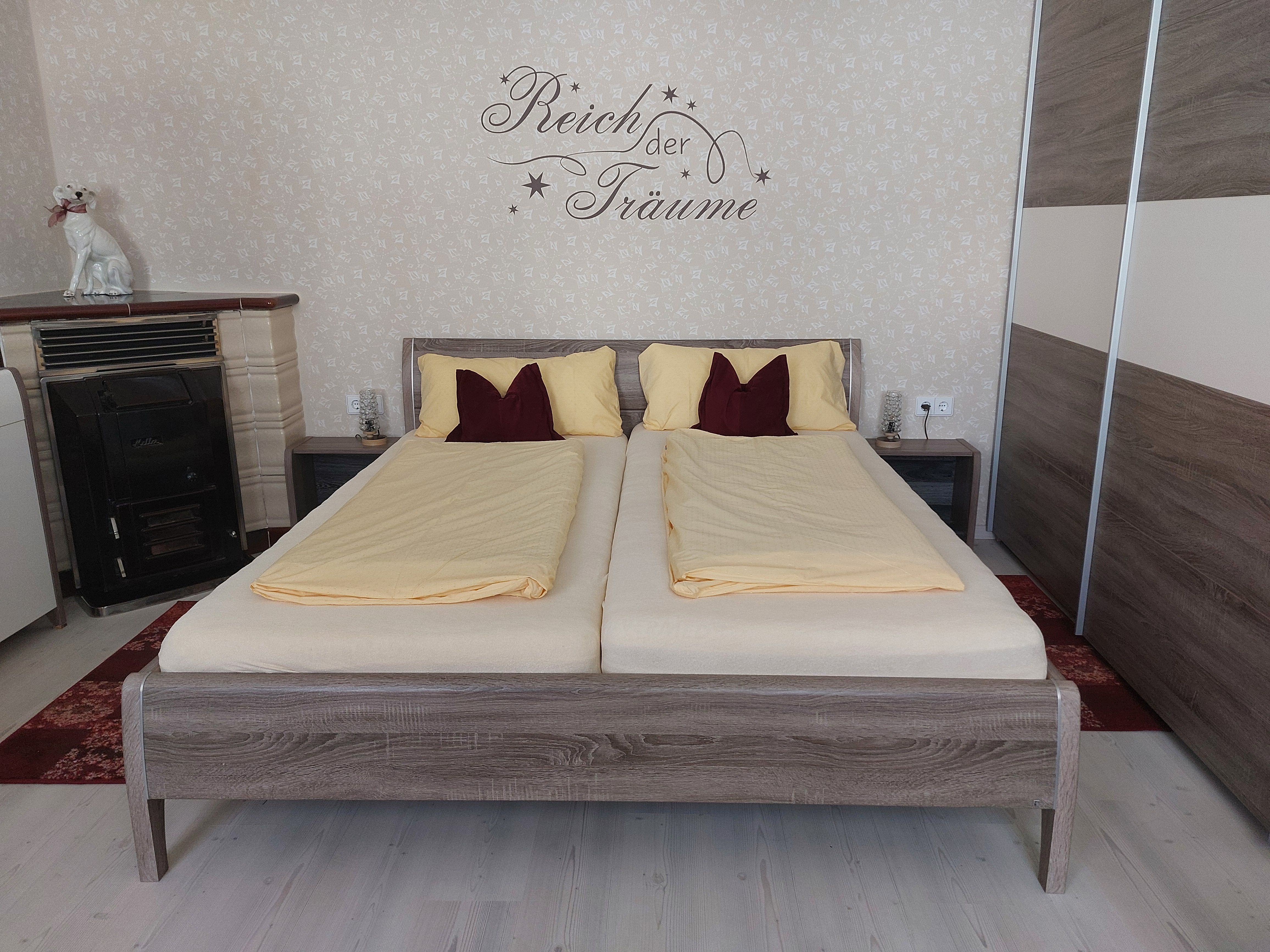 Spálňa s manželskou posteľou, žltou posteľnou bielizňou a nápisom na stene. Vidieť možno aj kachle a sochu psa.