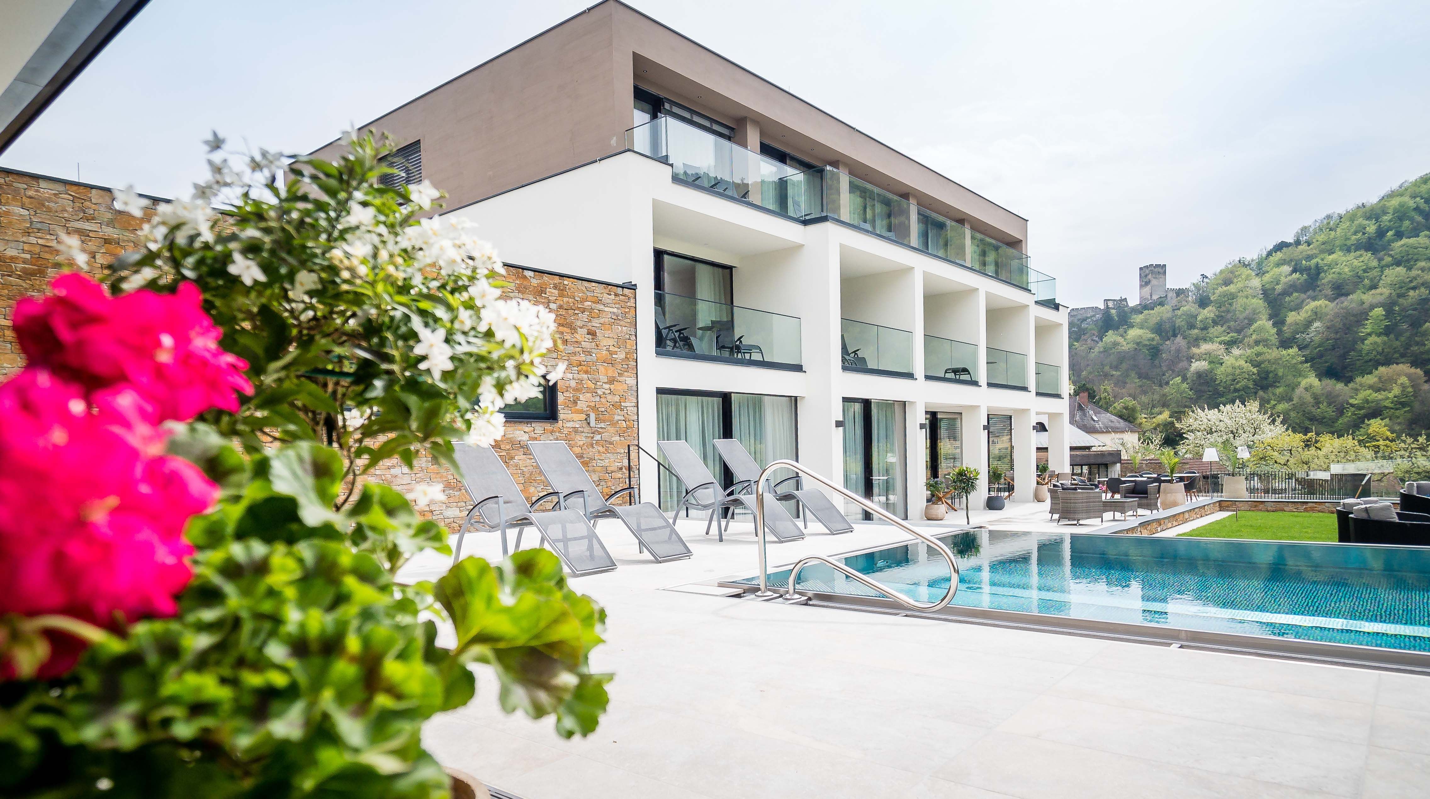 Moderná budova s bazénom a ležadlami, obklopená zelenou krajinou a hradom v pozadí.