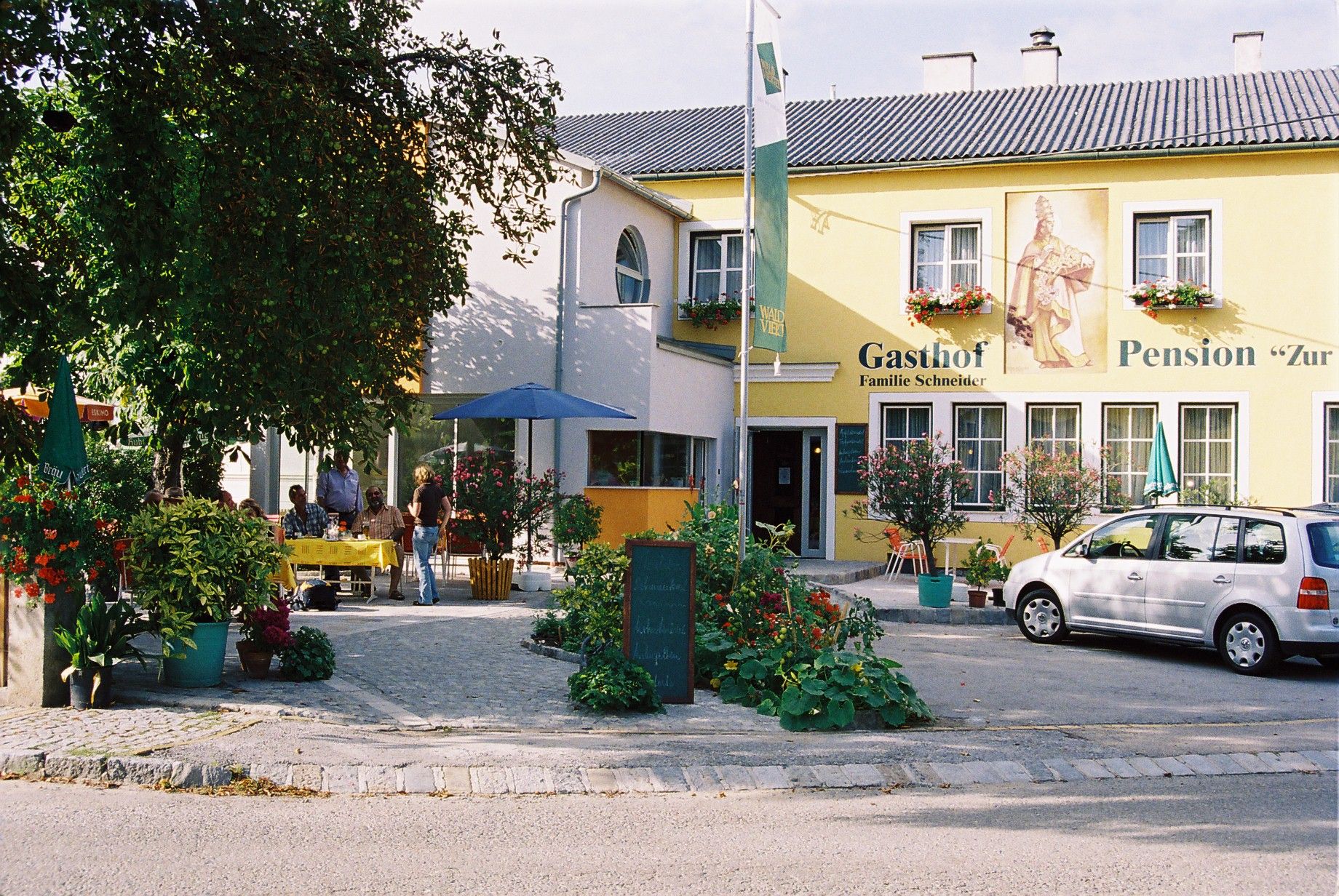 Žltá budova s označením "Gasthof-Pension Zur Hammerschmiede". Pred budovou sú stoly, stoličky a auto.