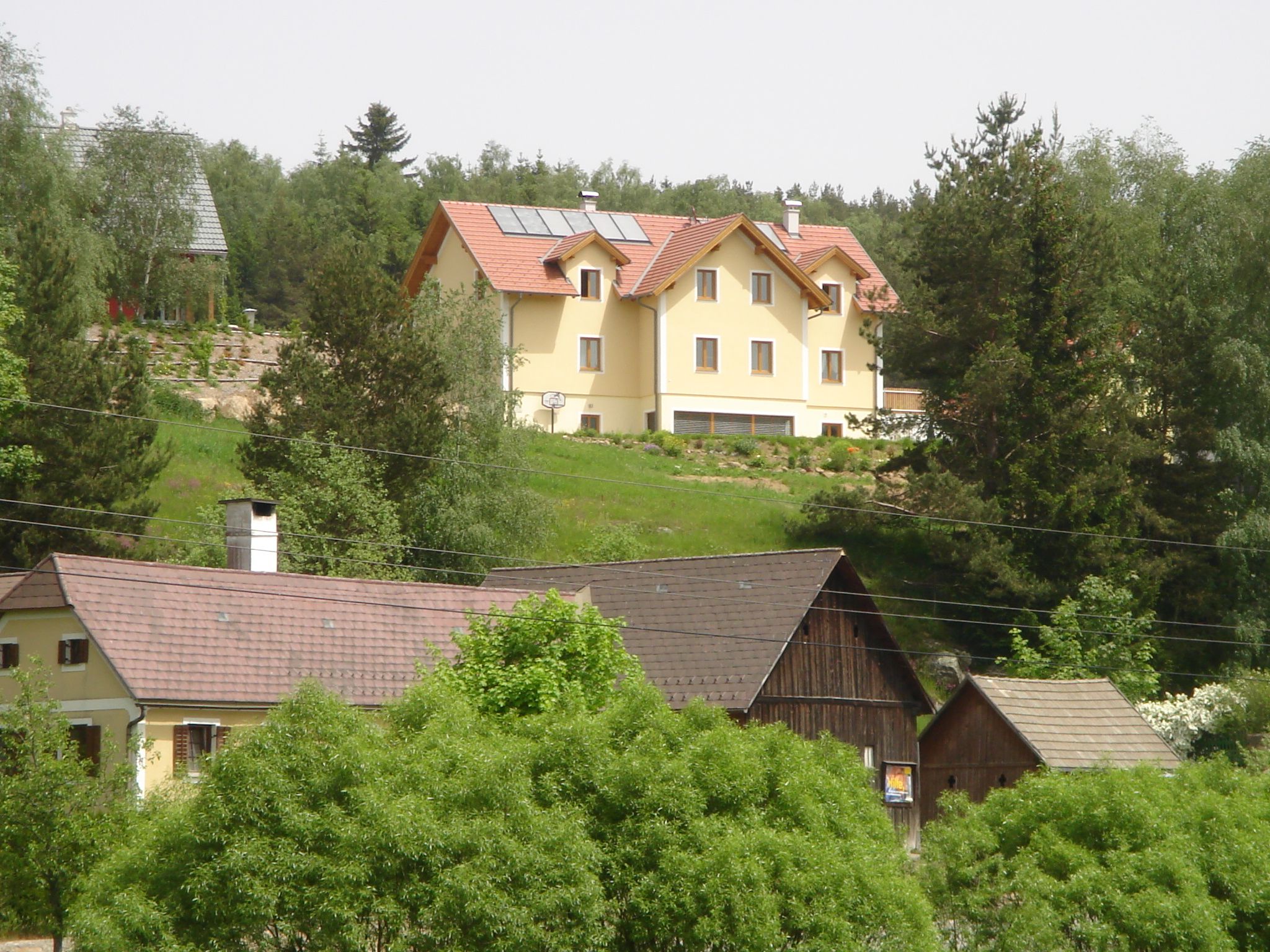 Veľký žltý dom so solárnymi panelmi na streche stojí na kopci, obklopený stromami a menšími budovami v popredí.