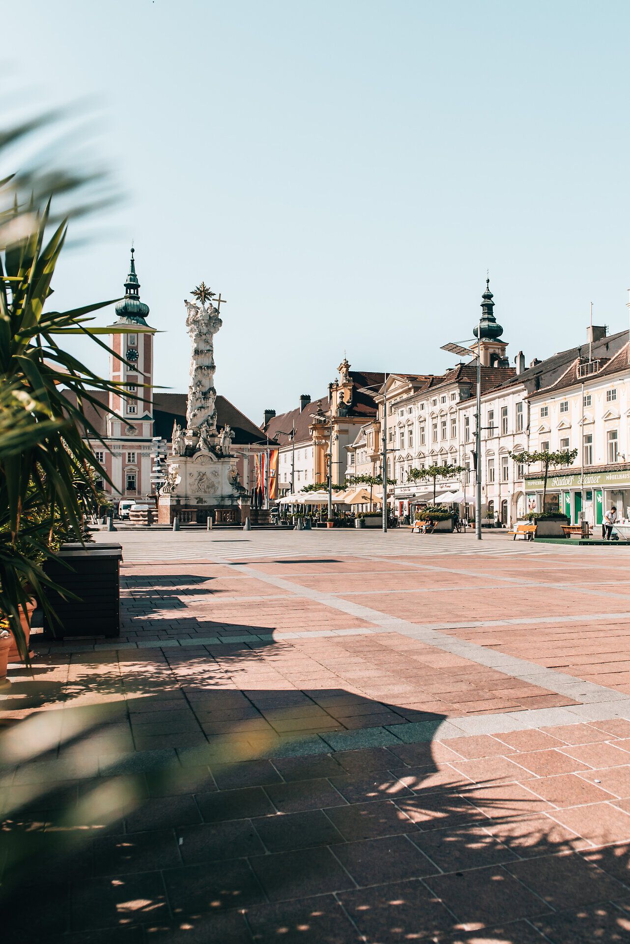 Die sonnendurchflutete Platzlandschaft lädt zum Verweilen ein, während die historischen Gebäude im Hintergrund eine charmante Kulisse bieten. Palmen und bunte Blumen verleihen dem Ort eine lebendige Atmosphäre, perfekt für entspannte Sommertage.