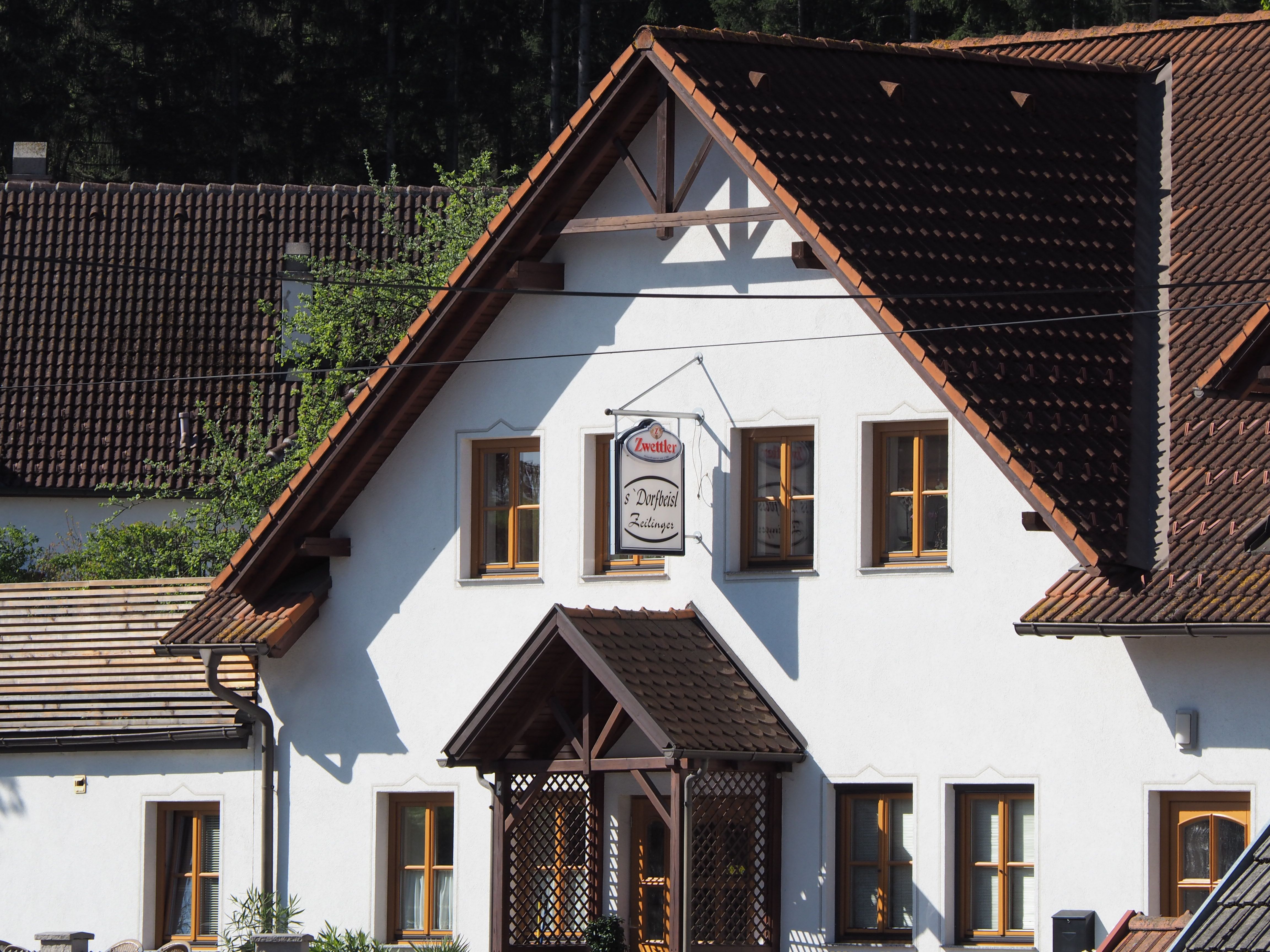 Tradičná budova s bielymi stenami a hnedými škridlami. Na fasáde visí nápis "Zwettler Dorfwirt Zeilinger".