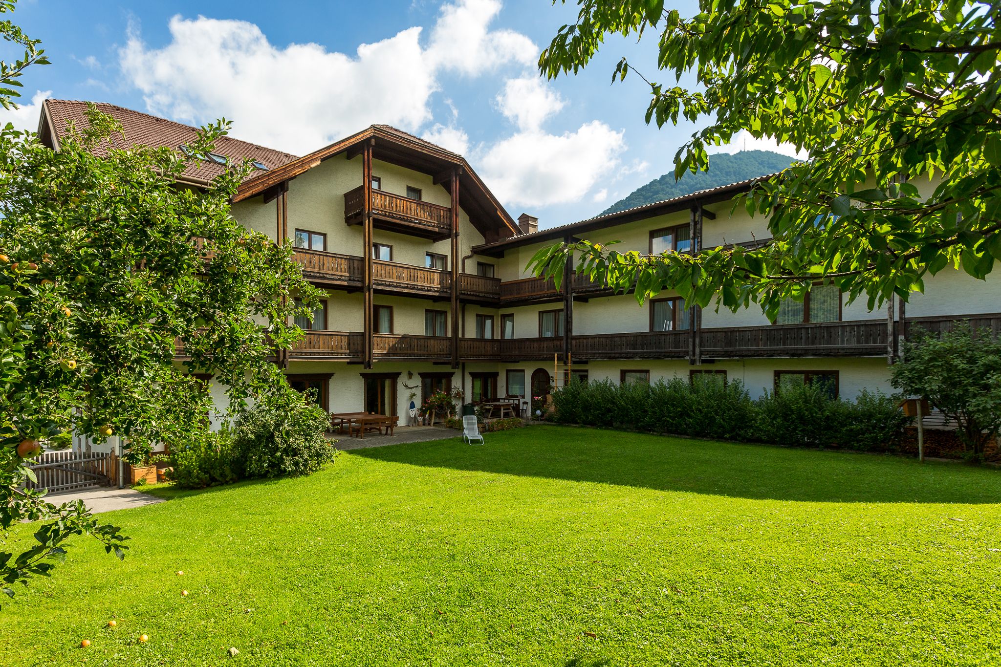 Tradičný hotel s drevenými balkónmi a udržiavanou záhradou v popredí, obklopený stromami a horami v pozadí.