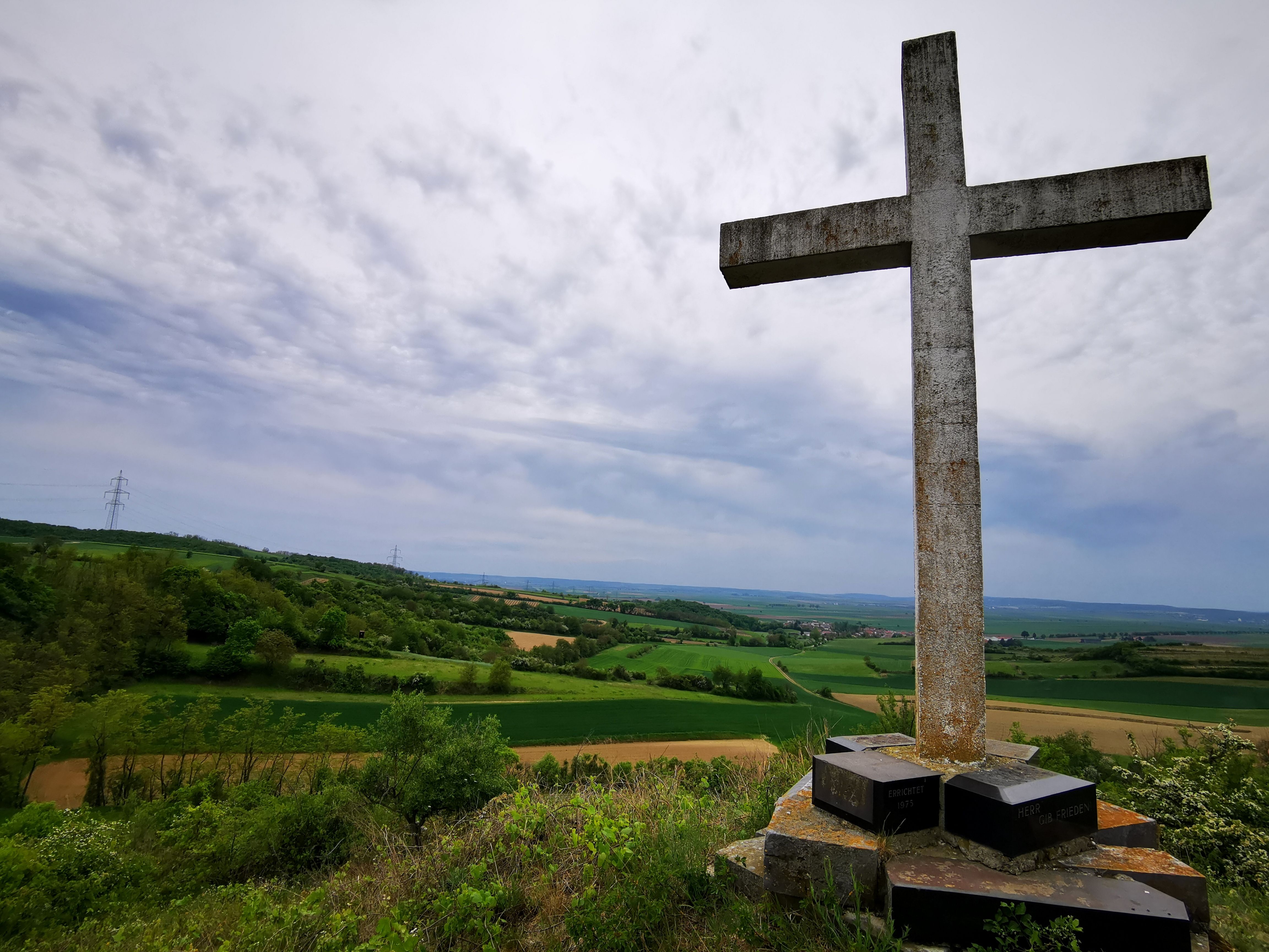 Veľký kamenný kríž na kopci s výhľadom na zelenú krajinu a zamračenú oblohu.