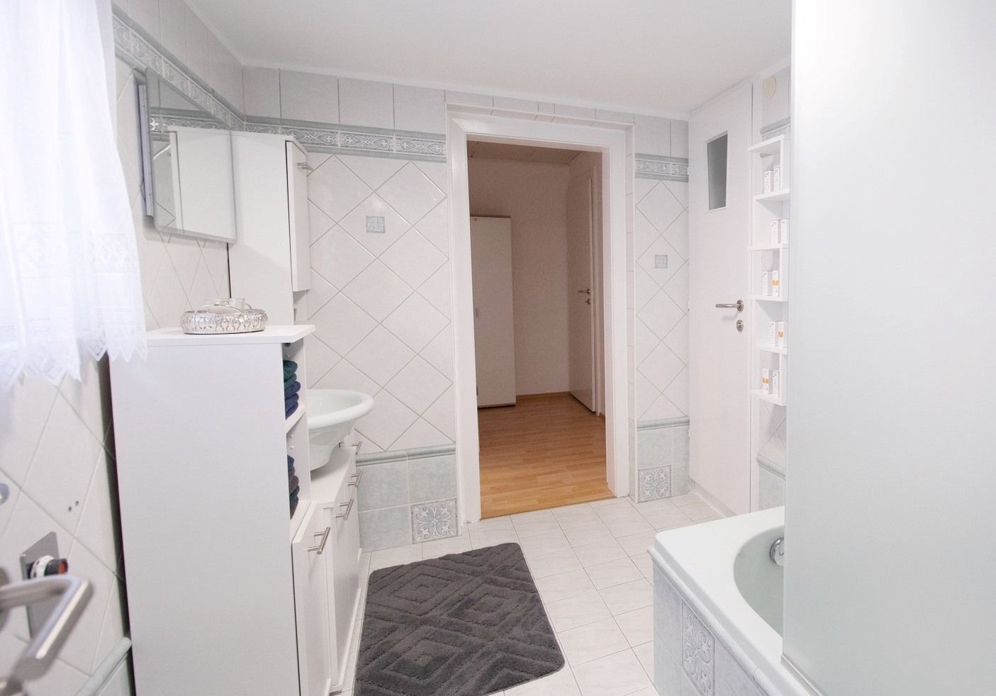 Moderná kúpeľňa s bielymi obkladačkami, vaňou, umývadlom a skrinkou. Dvere vedú do chodby s drevenou podlahou.