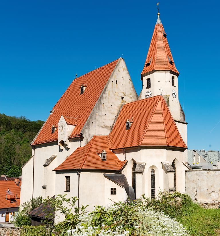 Edlitzský opevnený kostol s mnohými bočnými časťami, červenými škridlovými strechami, špicatou vežou a modrou oblohou.
