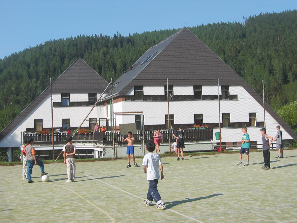 Deti hrajú futbal na športovom ihrisku pred hotelom v zalesnenej oblasti.