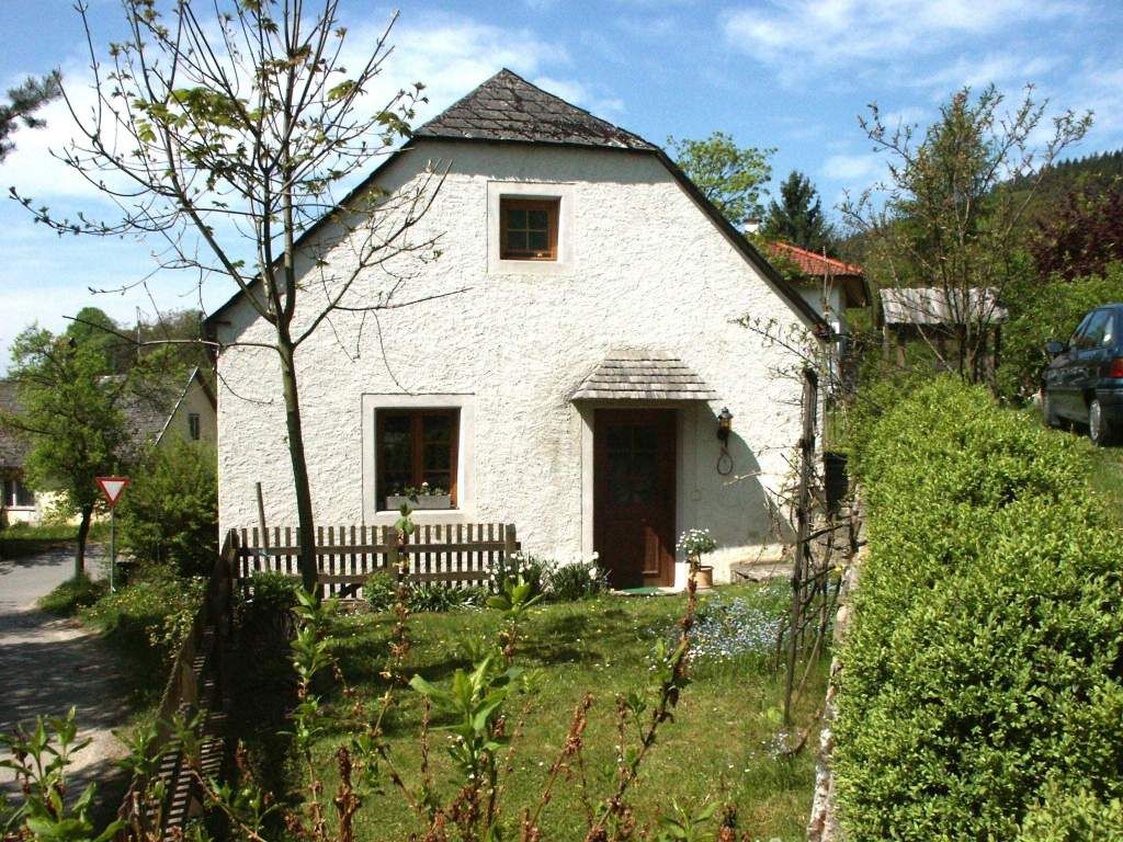 Malý biely rekreačný dom so záhradou a stromami, obklopený zelenou krajinou.