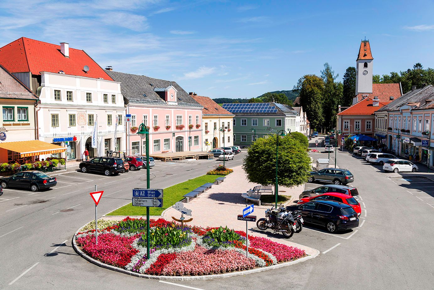 Hlavné námestie Aspang Markt s farebnými kvetinovými záhonmi, zaparkovanými autami a historickými budovami.
