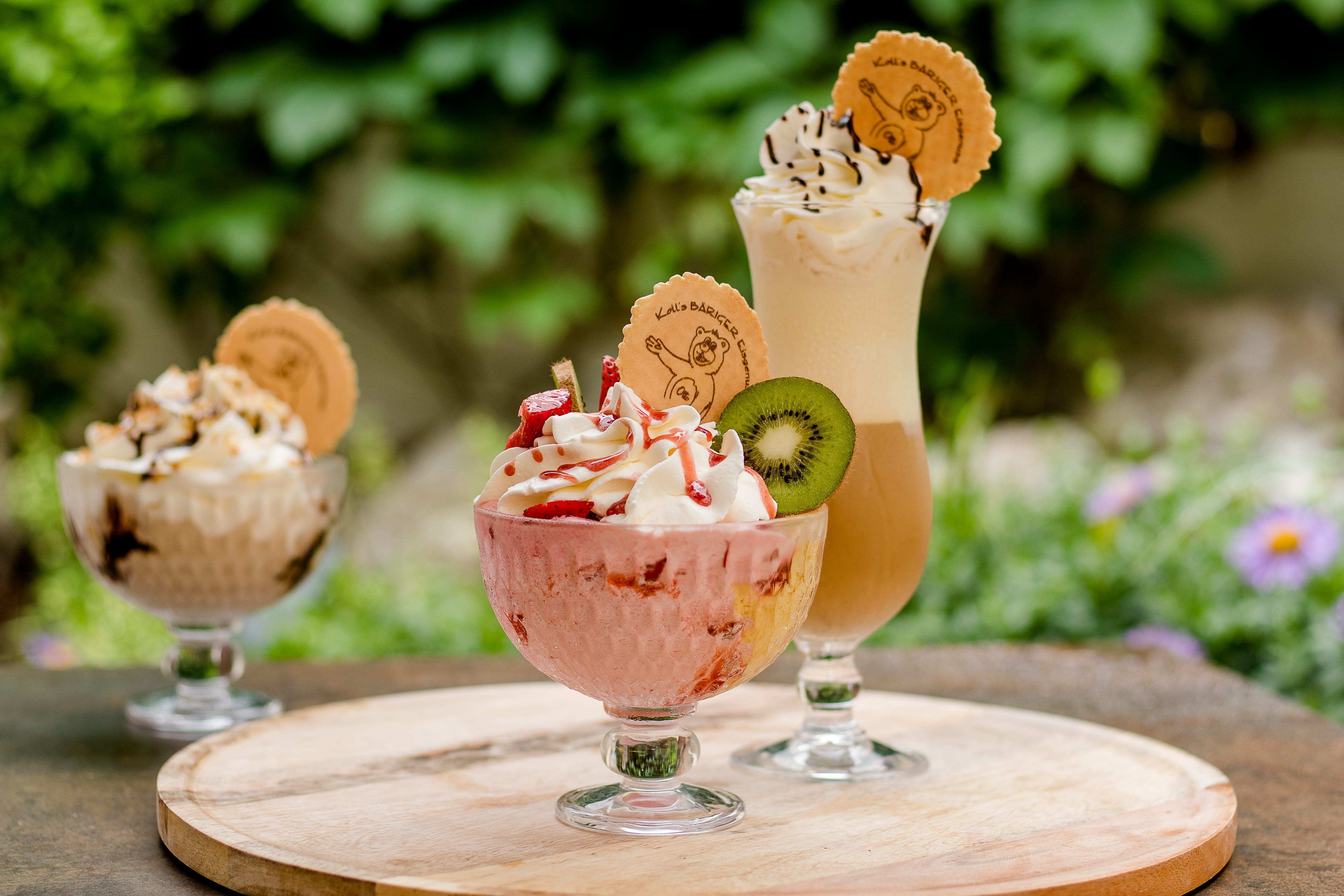 Tri poháre so šľahačkou a dekoráciou na drevenom stole vonku.