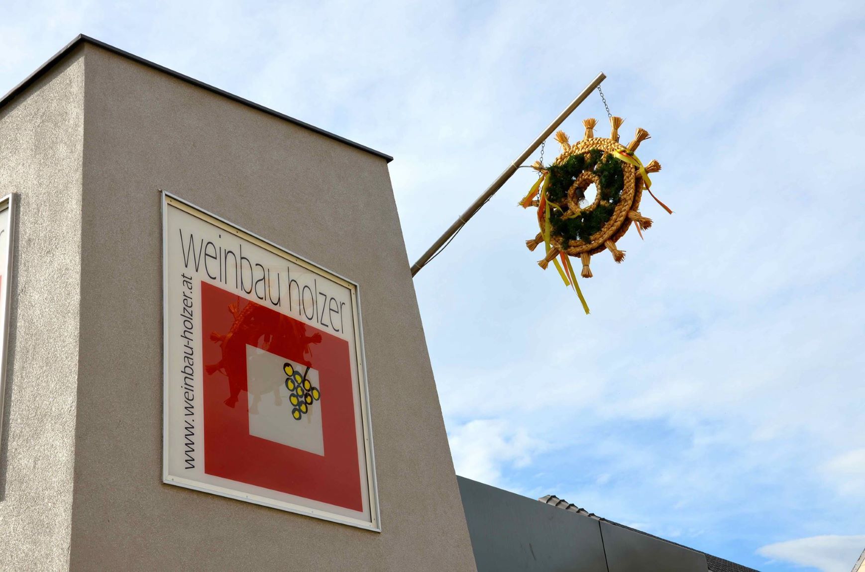Budova s nápisom "Weinbau Holzer" a ozdobným vencom na stĺpe.