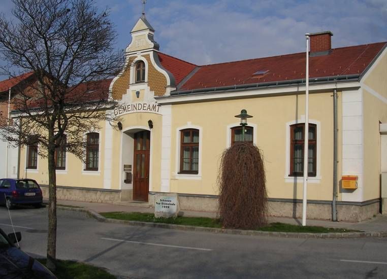Žltá budova s nápisom "Gemeindeamt", červenou strechou, stromom a lampášom pred ňou.