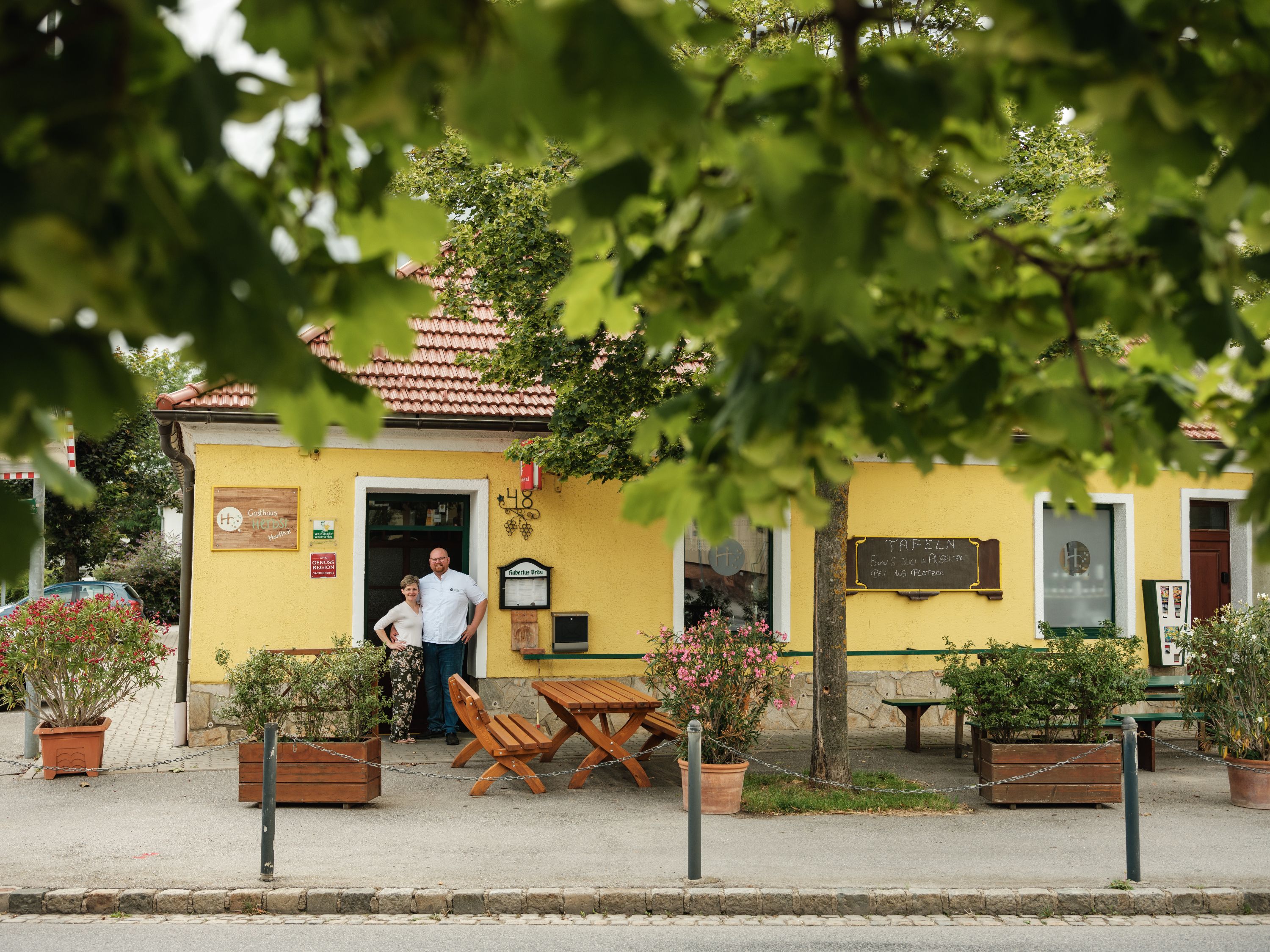 Žltý hostinec s červenou strechou, obklopený stromami. Pred vchodom stoja dvaja ľudia.