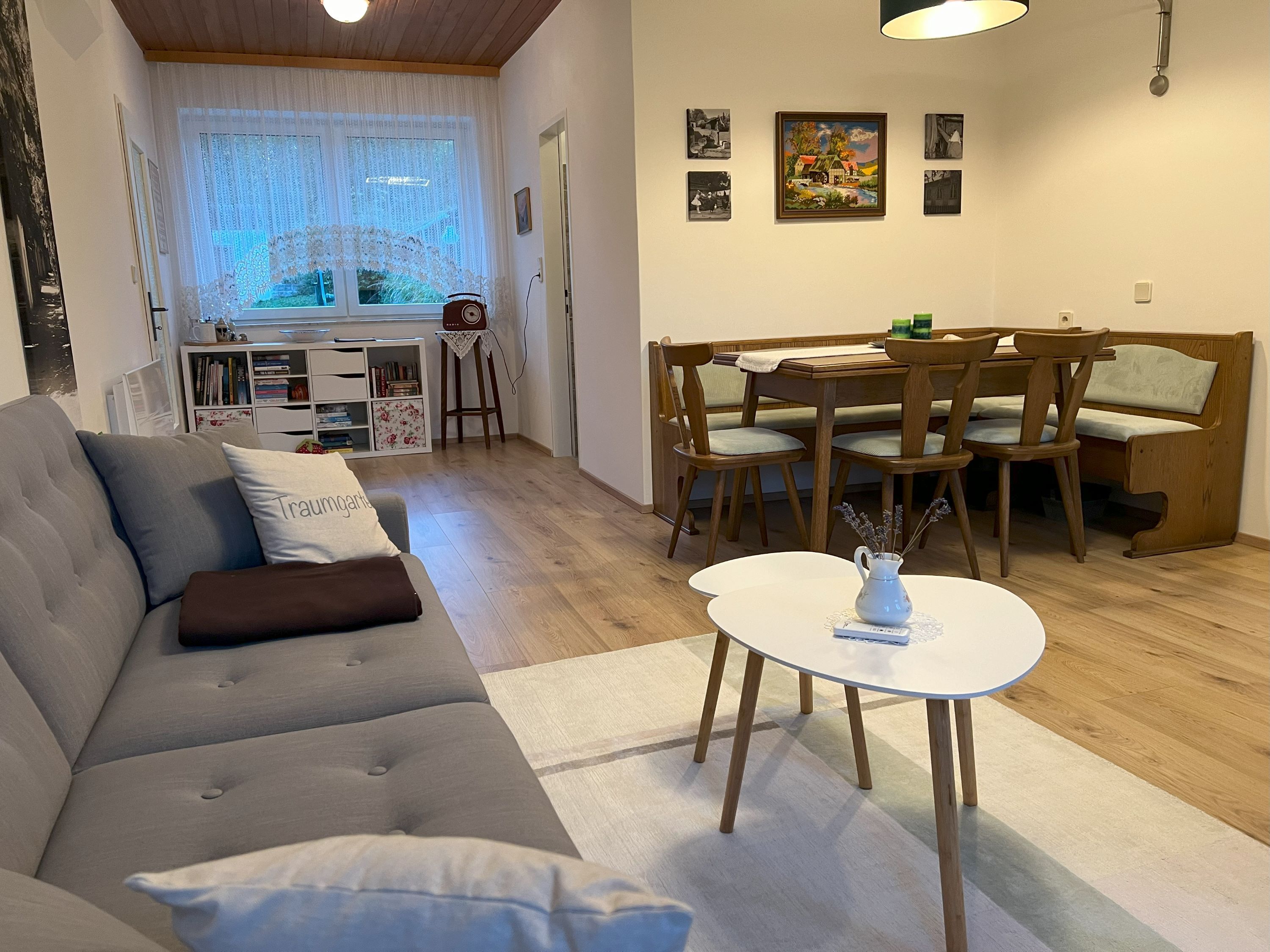 Útulná obývacia izba so sivou pohovkou, jedálenským stolom a stoličkami, policou na knihy a oknom so závesmi.