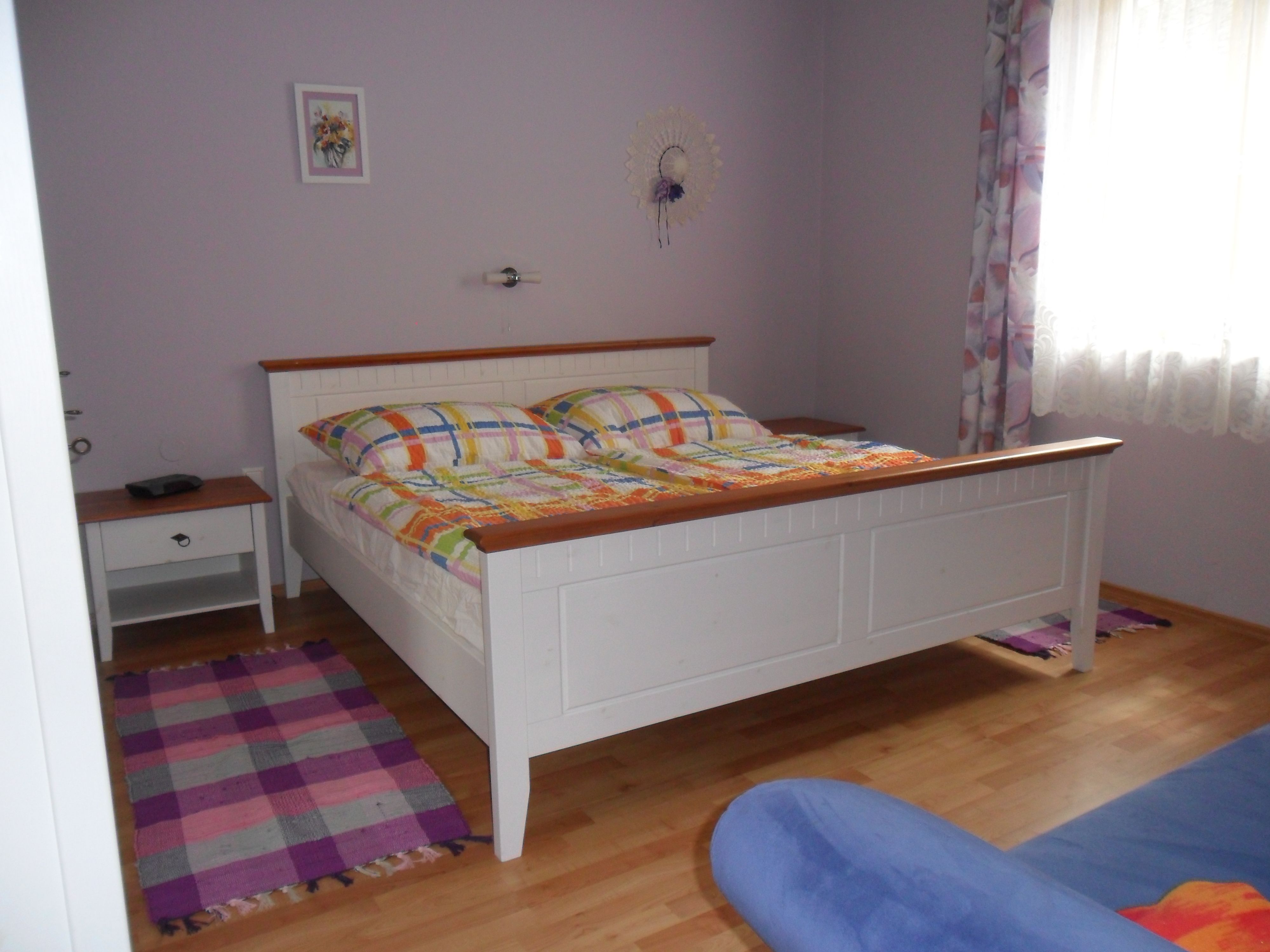 Spálňa s bielou posteľou, farebnými perinami, nočným stolíkom a oknom so závesmi.