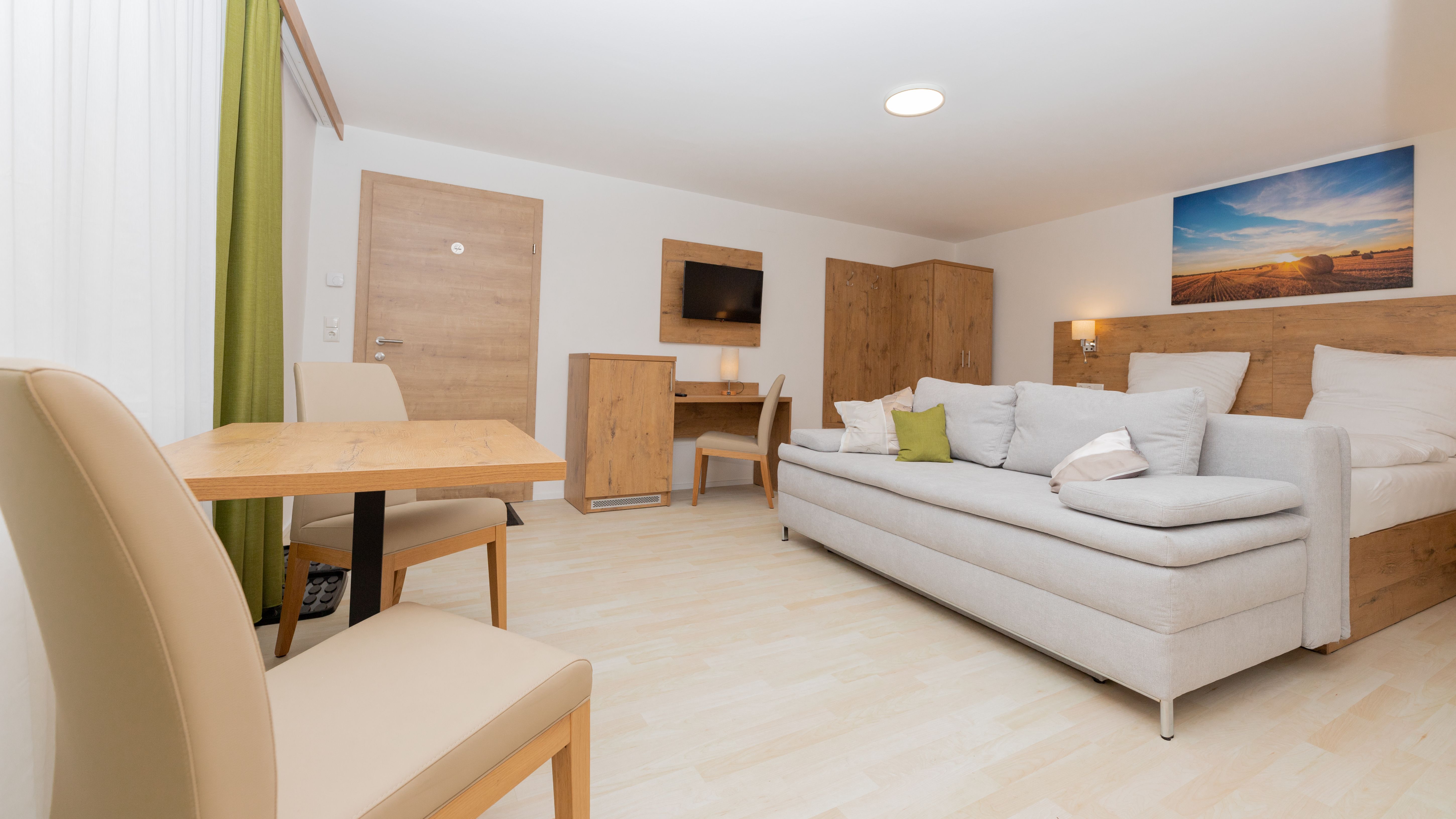 Moderná spálňa s rozkladacou pohovkou, stolom a stoličkami, dreveným nábytkom a obrazom krajiny na stene.