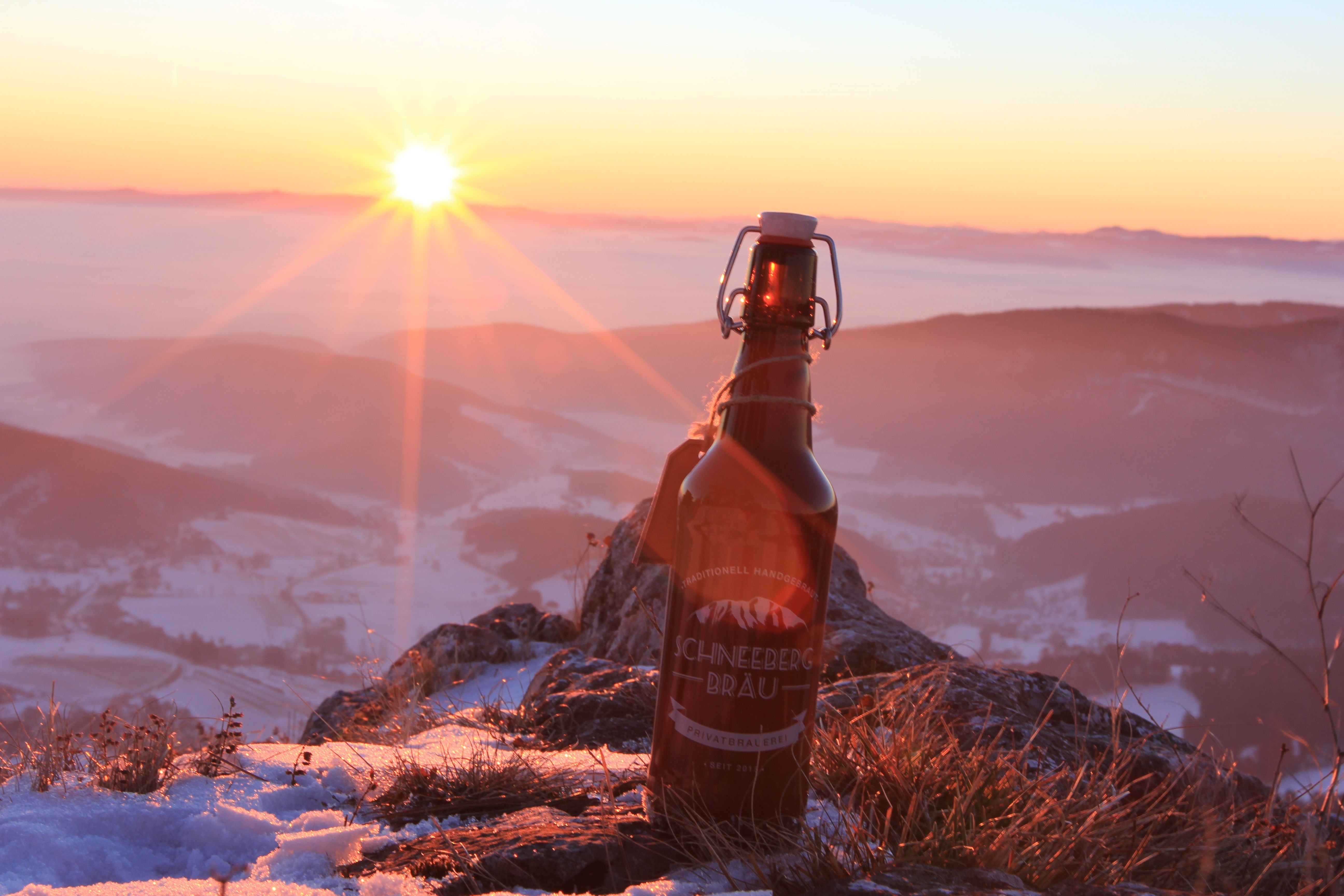 Fľaša piva Schneeberg Bräu stojí na zasneženej hore pri západe slnka.