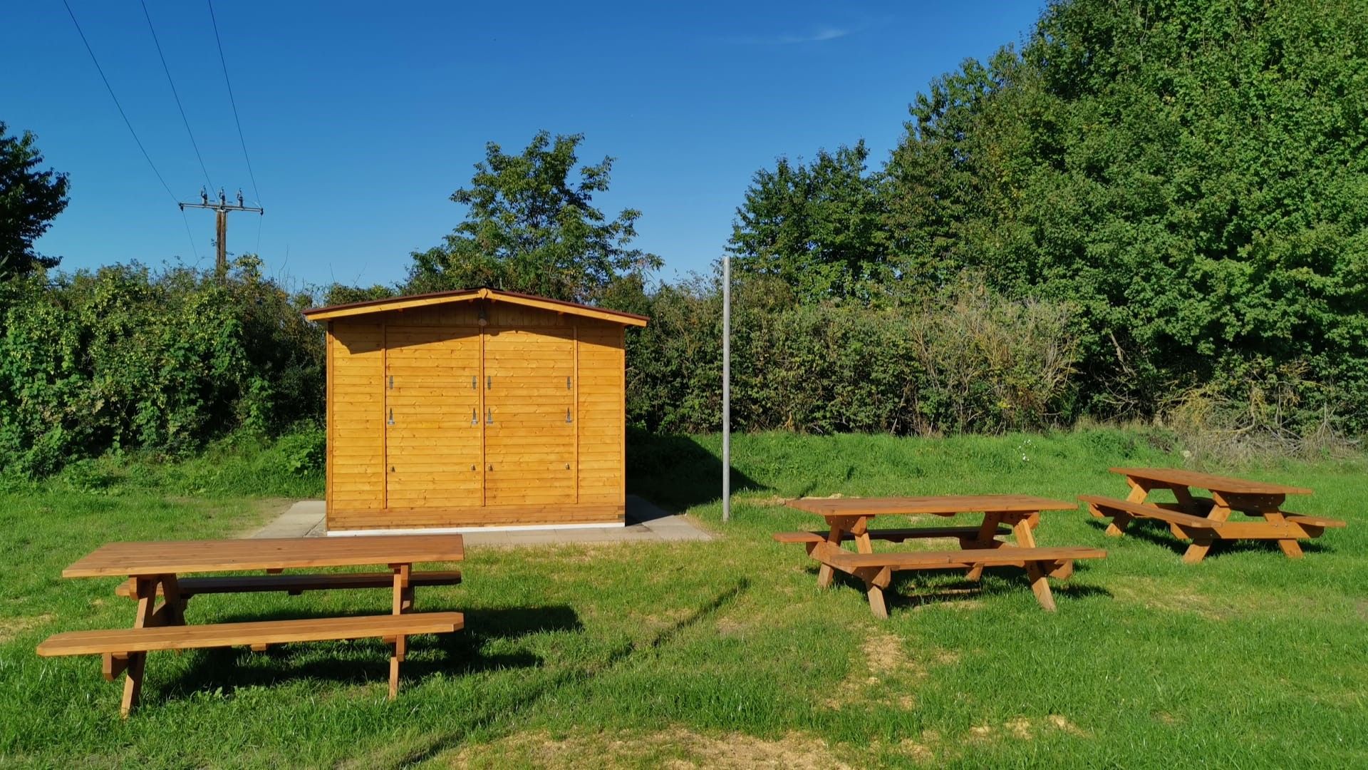 Drevená chatka s piknikovými stolmi na lúke, obklopená stromami a kríkmi pod modrou oblohou.