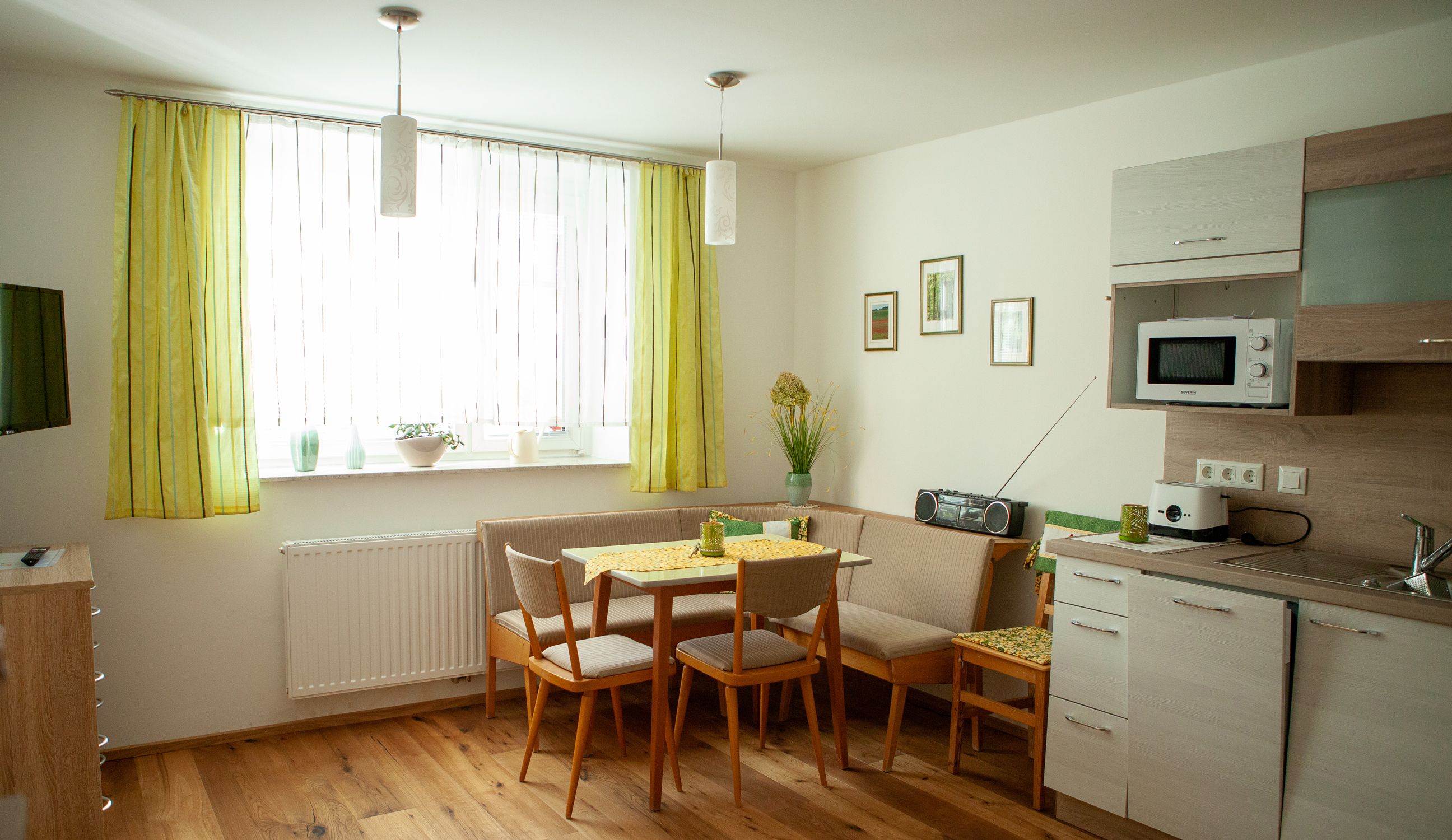 Útulná kuchyňa s jedálenským stolom, rohovou lavicou, kuchynskou linkou a zelenými závesmi.