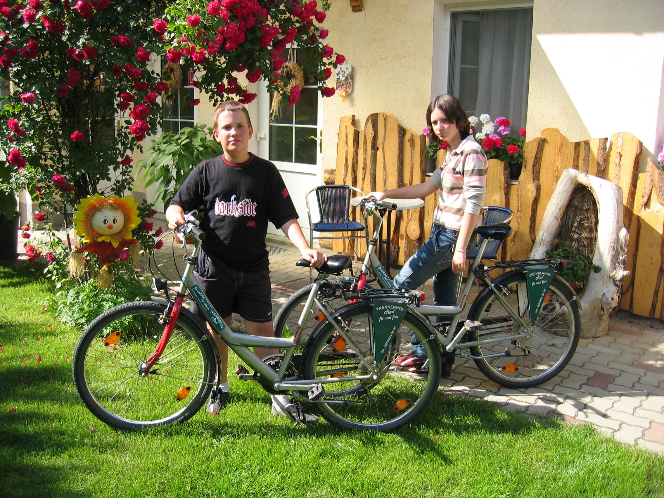 Dvaja ľudia s bicyklami pred domom s kvetmi a drevenými ozdobami.