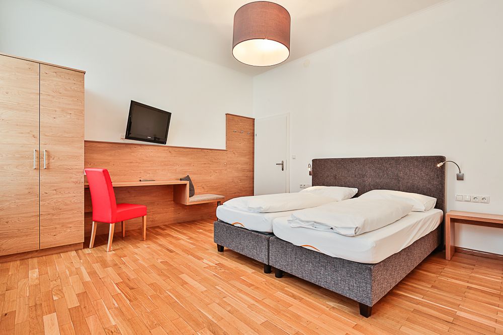 Hotelová izba s manželskou posteľou, červenou stoličkou, písacím stolom a televízorom.