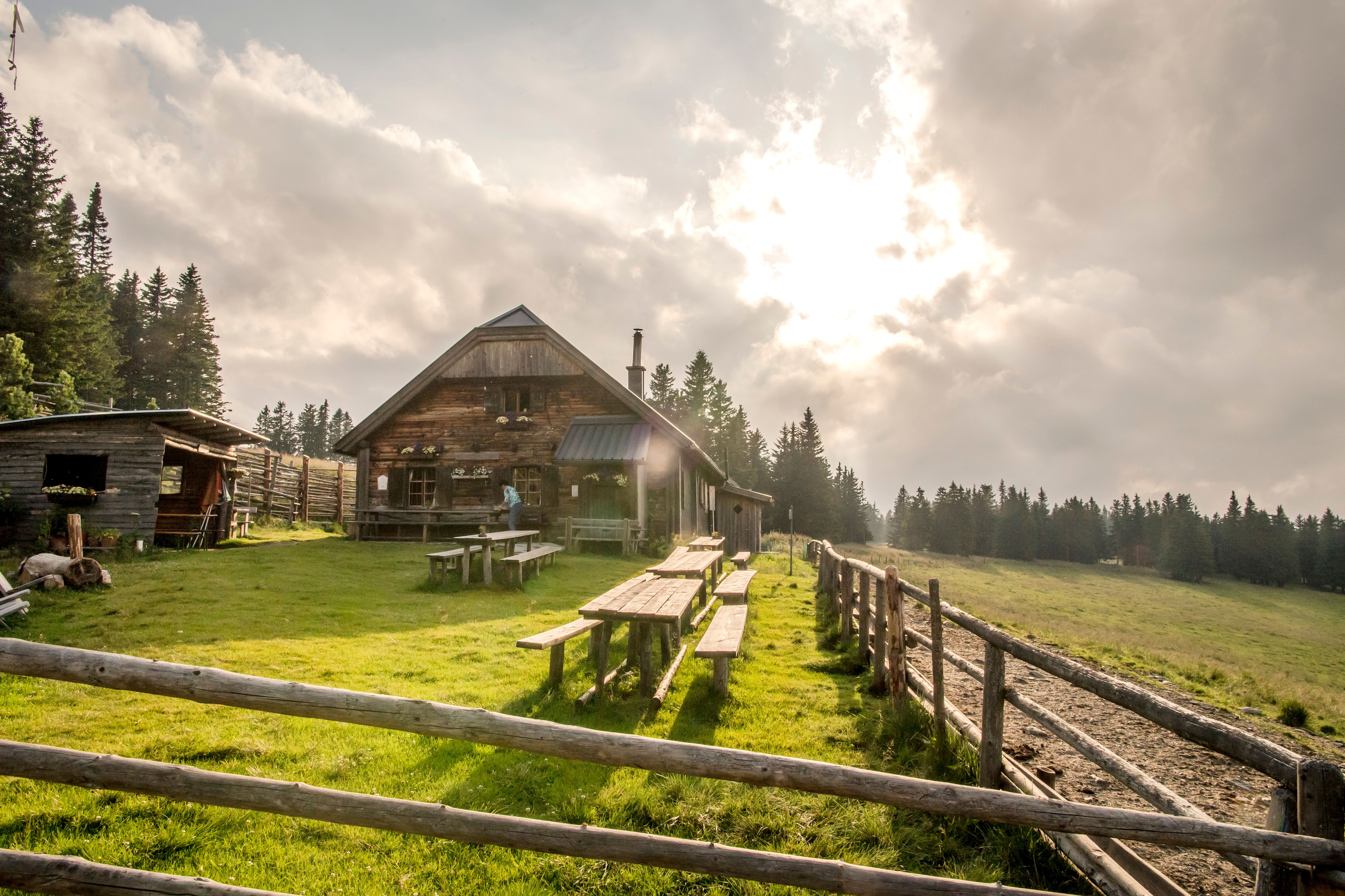 Alpská chata s drevenými stolmi na lúke, obklopená lesom, pod zamračenou oblohou.