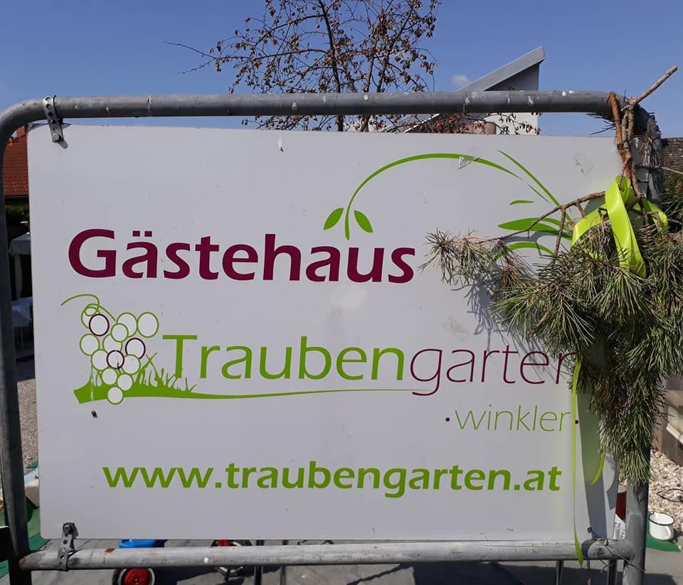 Tabuľka s nápisom "Gästehaus Traubengarten Winkler" a internetovou adresou, zdobená vetvičkami.