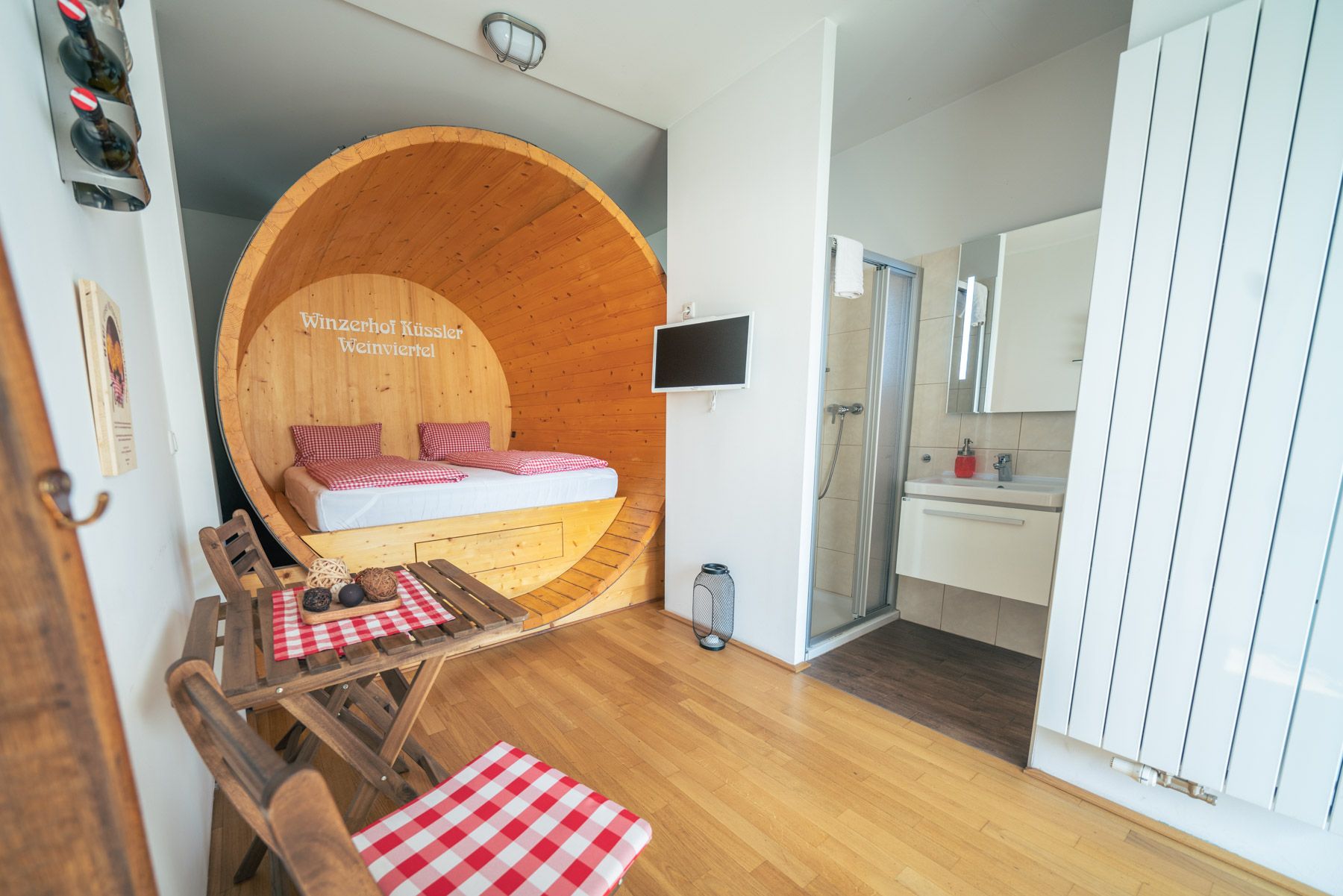 Pohľad do interiéru apartmánu s posteľou vo veľkom sude na víno, malým stolom so stoličkami a kúpeľňou.
