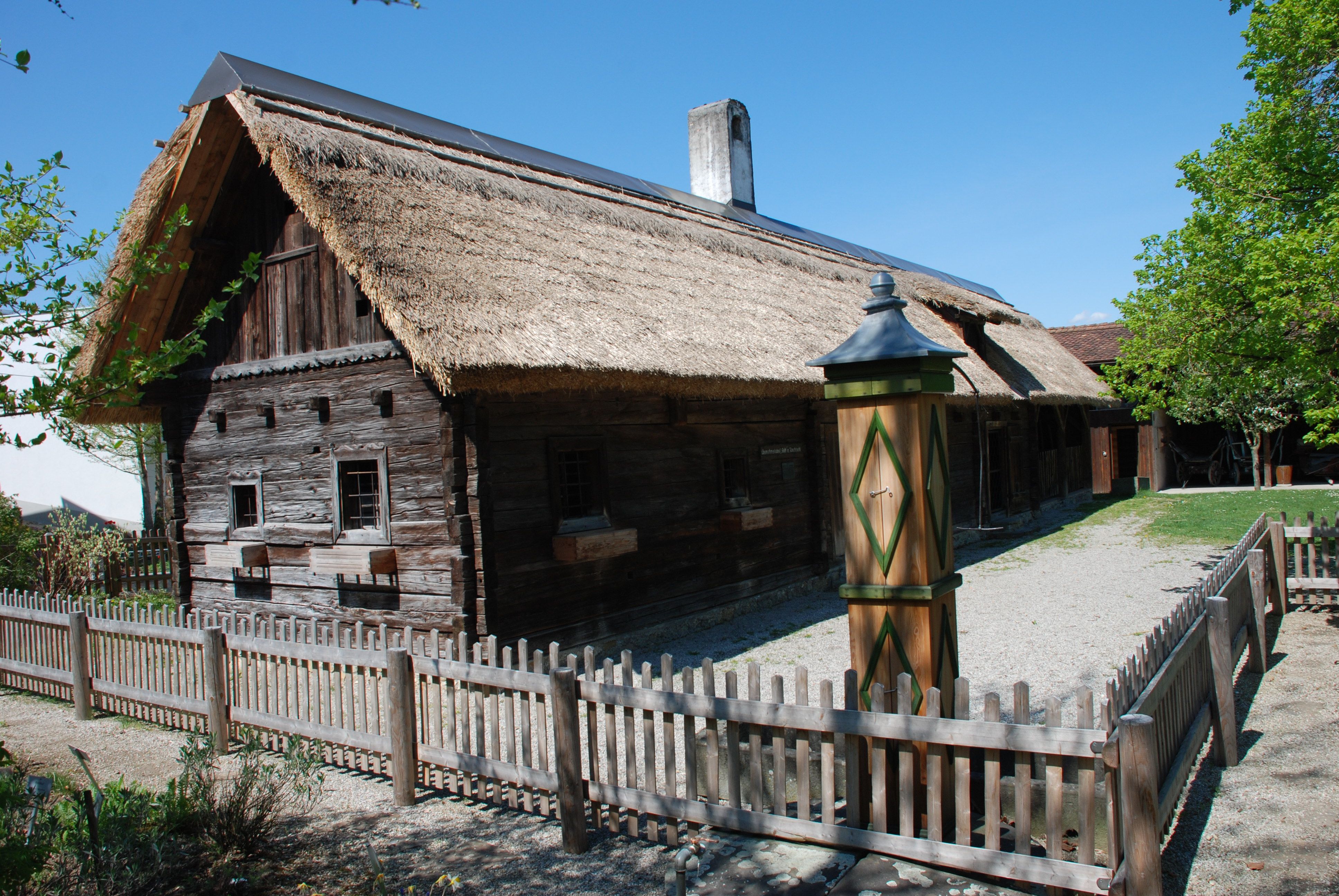 Tradičný drevený dom so slamenou strechou a dreveným plotom vo vidieckom prostredí.